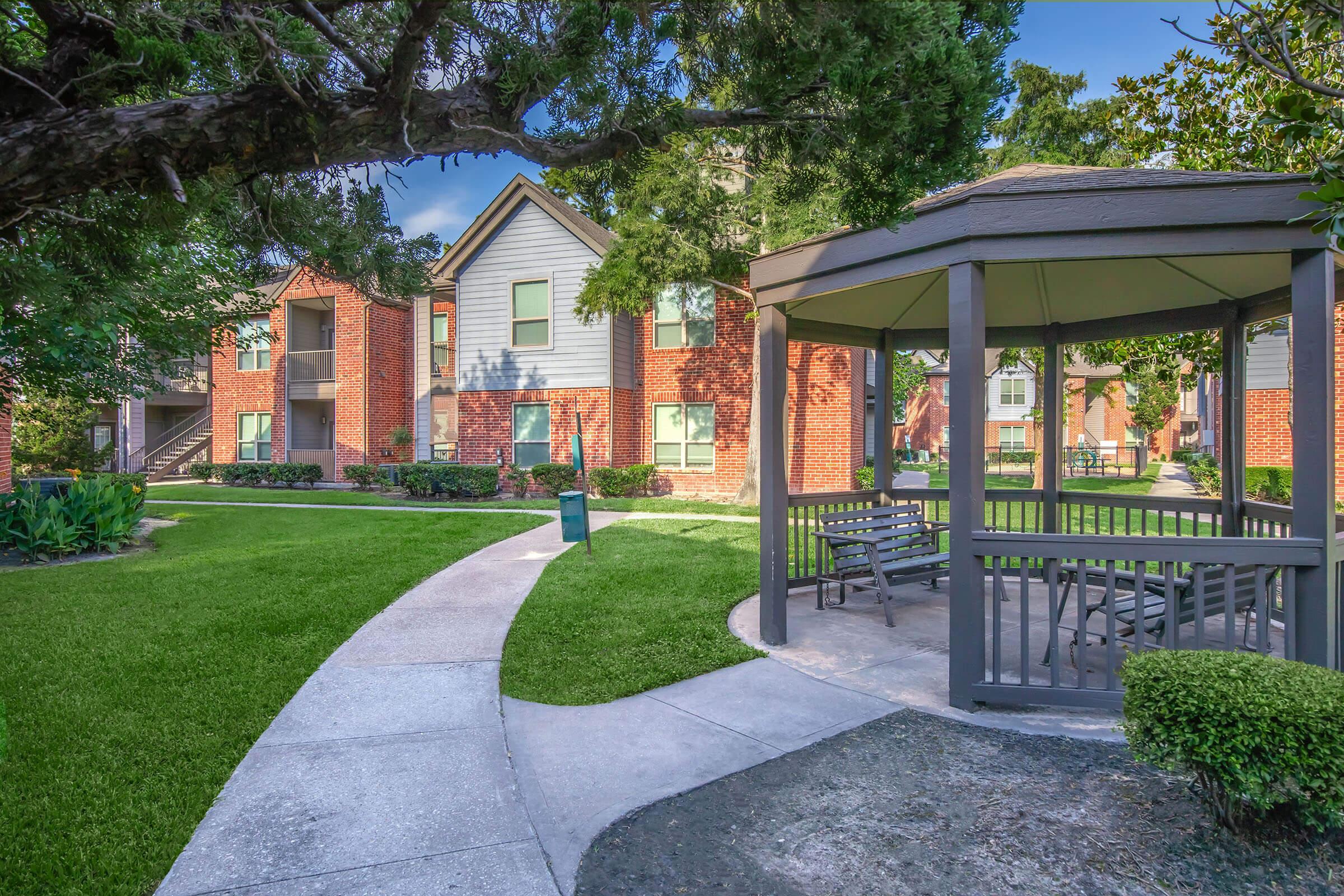 A well-maintained apartment complex featuring two-story brick buildings surrounded by lush green lawns and trees. A gazebo with benches sits in the foreground, offering a shaded seating area, while a curved walkway connects the gazebo to the apartment buildings. Clear blue skies are visible in the background.