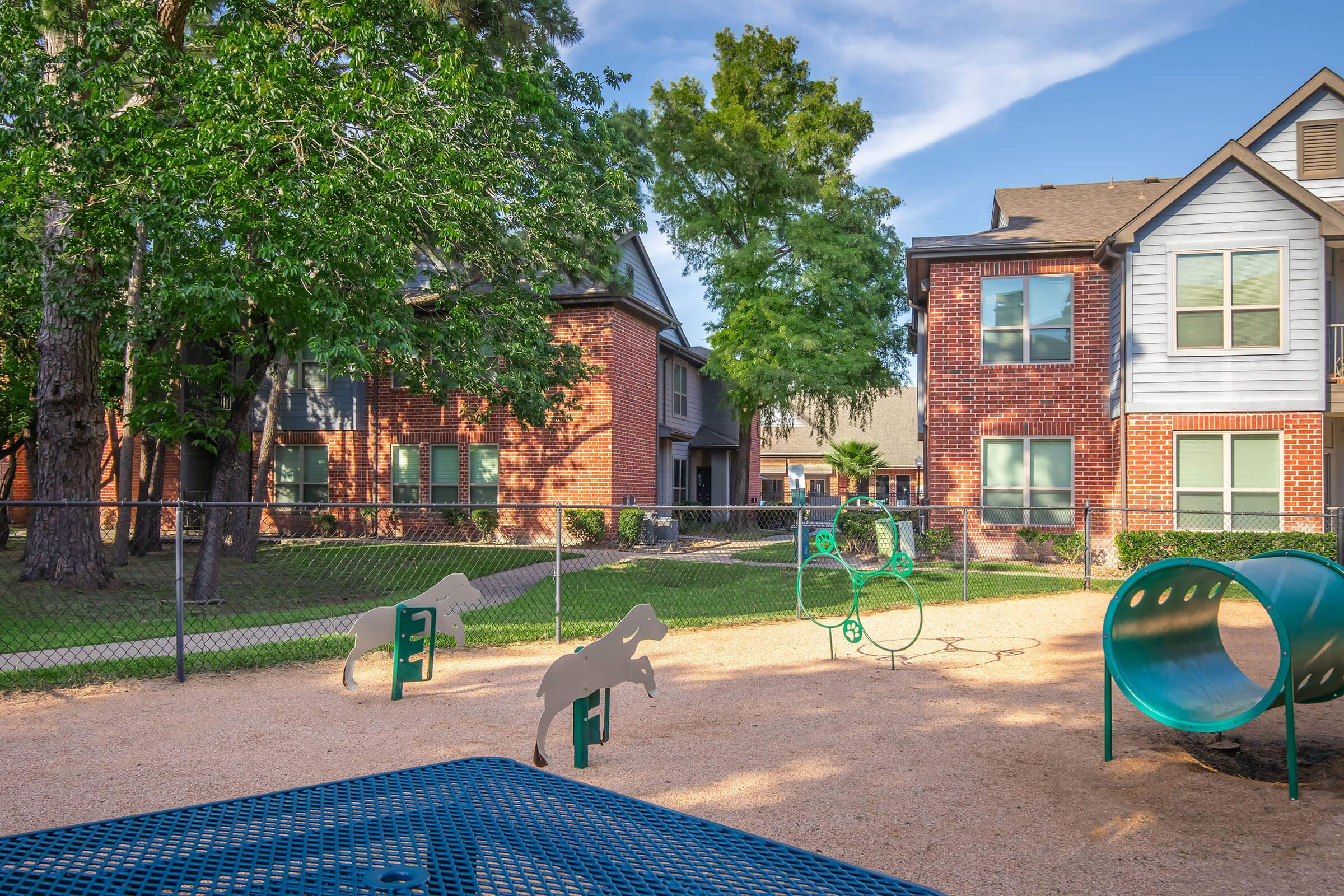 A playground area featuring exercise equipment and play structures, surrounded by a fenced yard. The background includes residential buildings with brick exteriors and green trees, providing a serene atmosphere. The ground is covered in gravel, creating a safe play environment for children and pets.