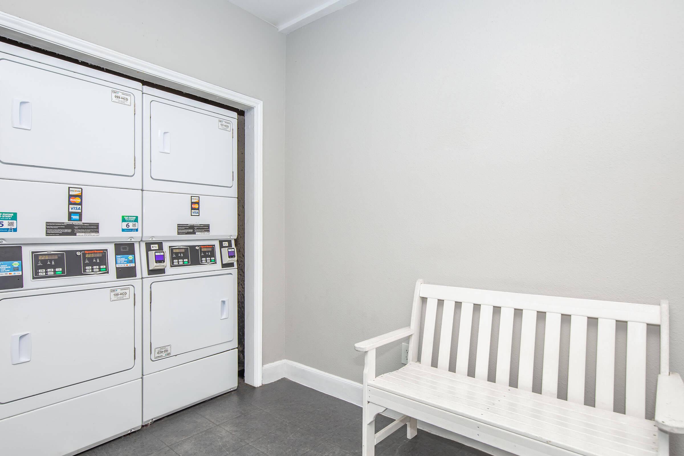 A clean, well-lit laundry room featuring a row of stacked washing machines and dryers against a gray wall. To the side, there is a white wooden bench for seating. The floor is dark, creating a modern and spacious feel.