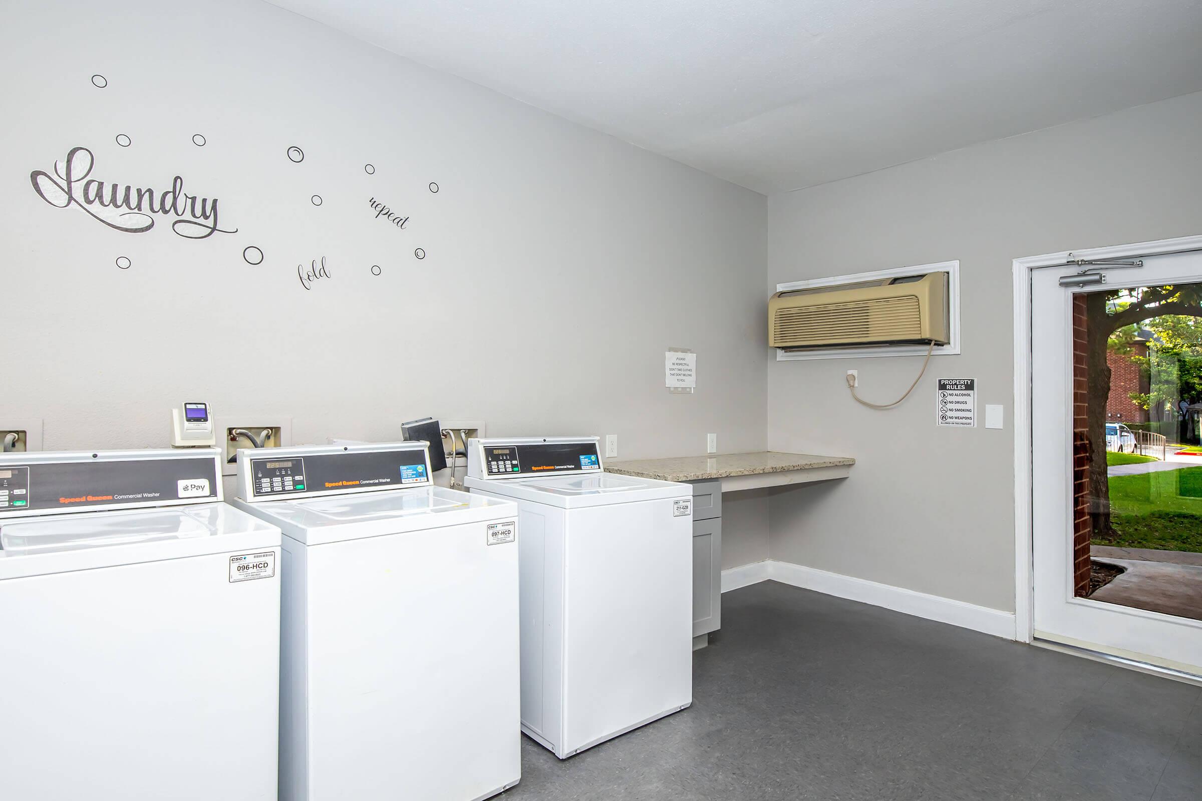 A clean and modern laundry room featuring several white washing machines and a countertop. The walls are painted grey with decorative text reading "Laundry" and small dots. An air conditioning unit is mounted on the wall, and a door on the right leads to an outdoor area. Natural light enters through the door.