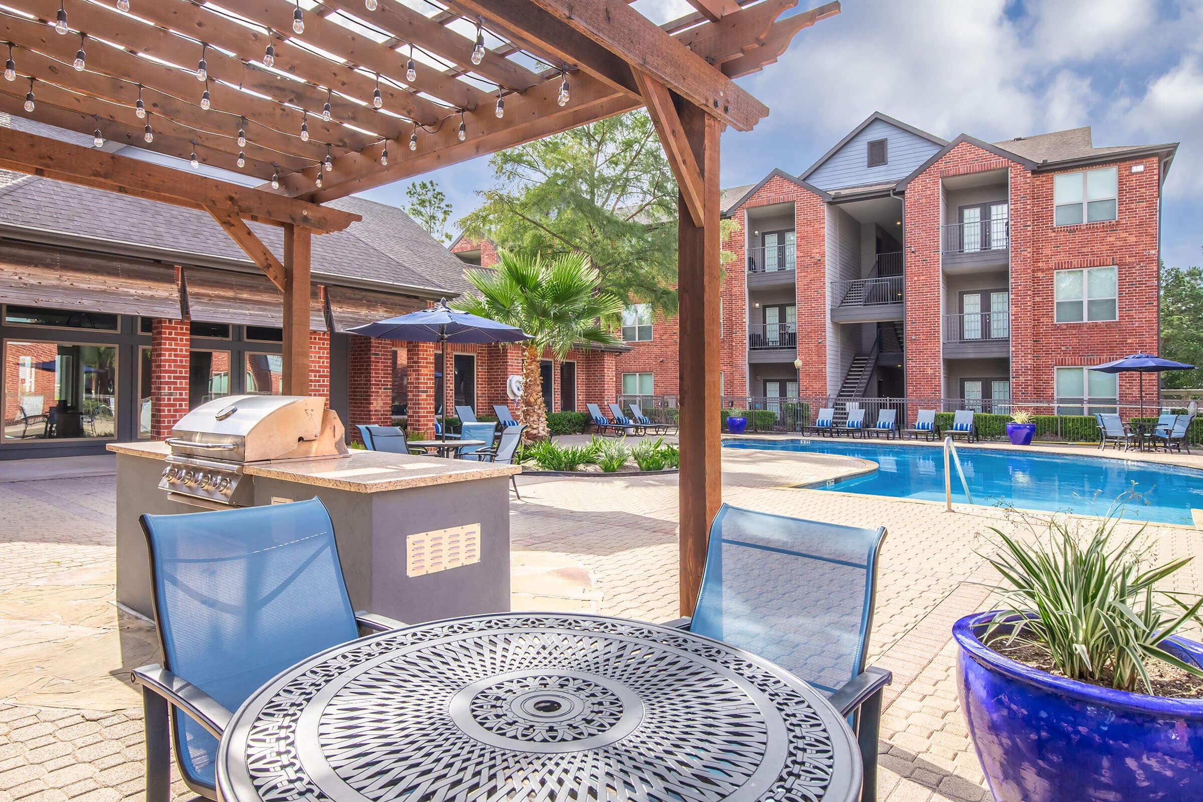 Outdoor pool area with lounge chairs, a shaded seating area featuring a table and blue chairs, a grill, and landscaped greenery. Red brick apartment buildings are in the background under a partly cloudy sky.