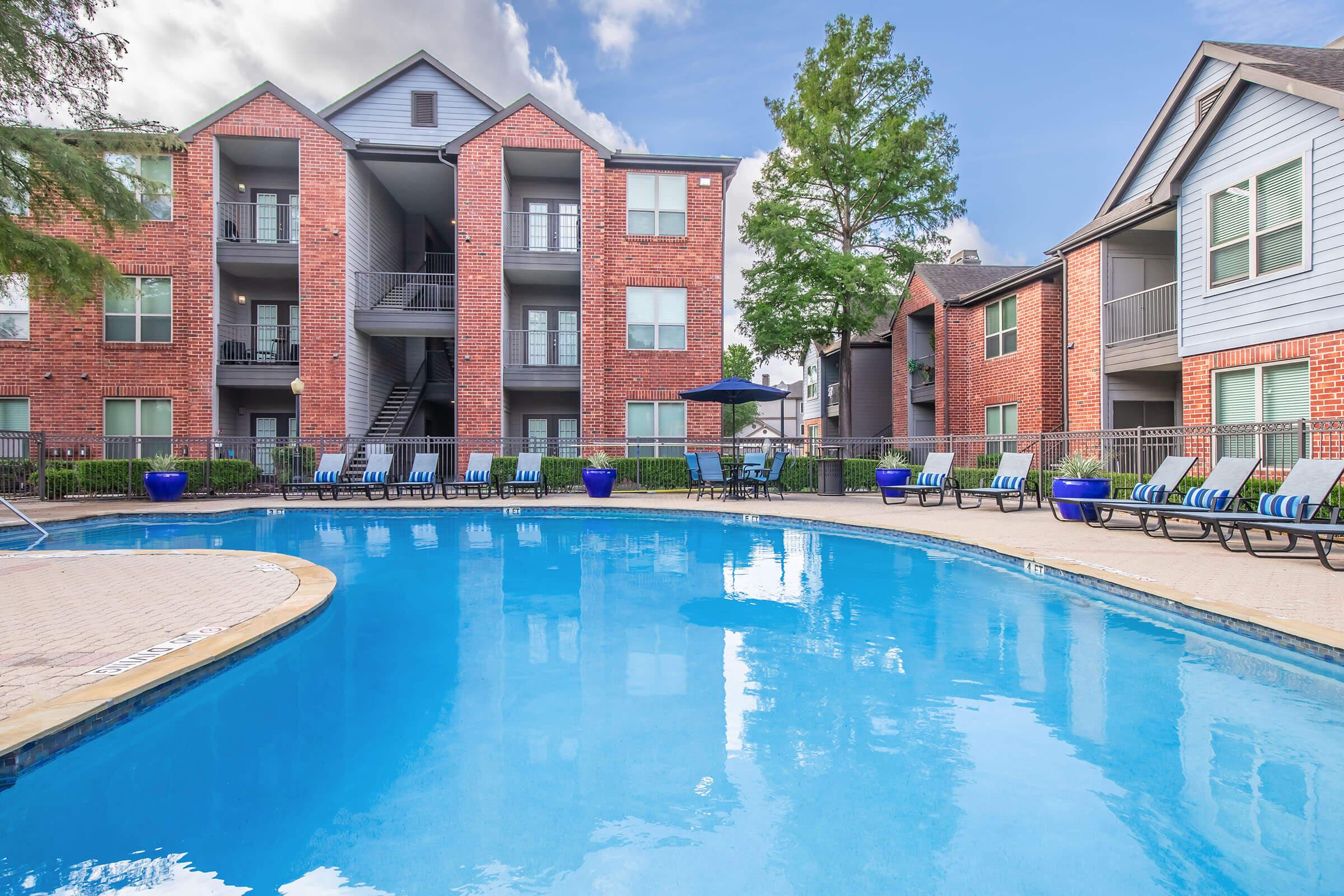 A clear blue swimming pool surrounded by lounge chairs and shaded areas, with several apartment buildings in the background. The scene is set under a partly cloudy sky, showcasing a relaxing outdoor environment.
