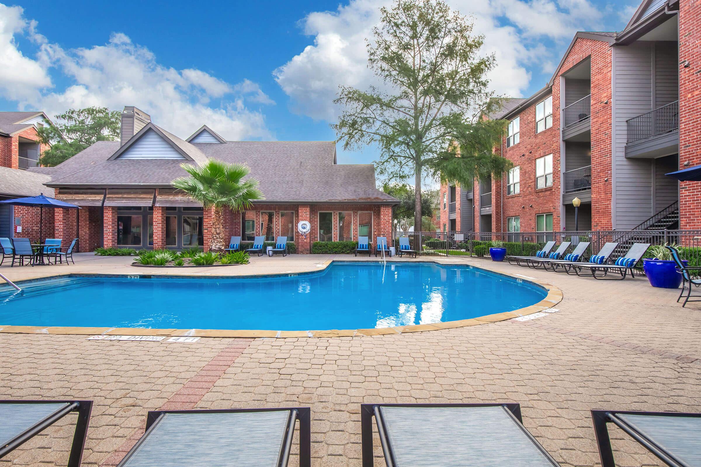 A serene swimming pool surrounded by lounge chairs and palm trees, set within a well-maintained apartment complex. The buildings feature brick exteriors, and the sky is bright with fluffy clouds, creating a tranquil outdoor space for relaxation.