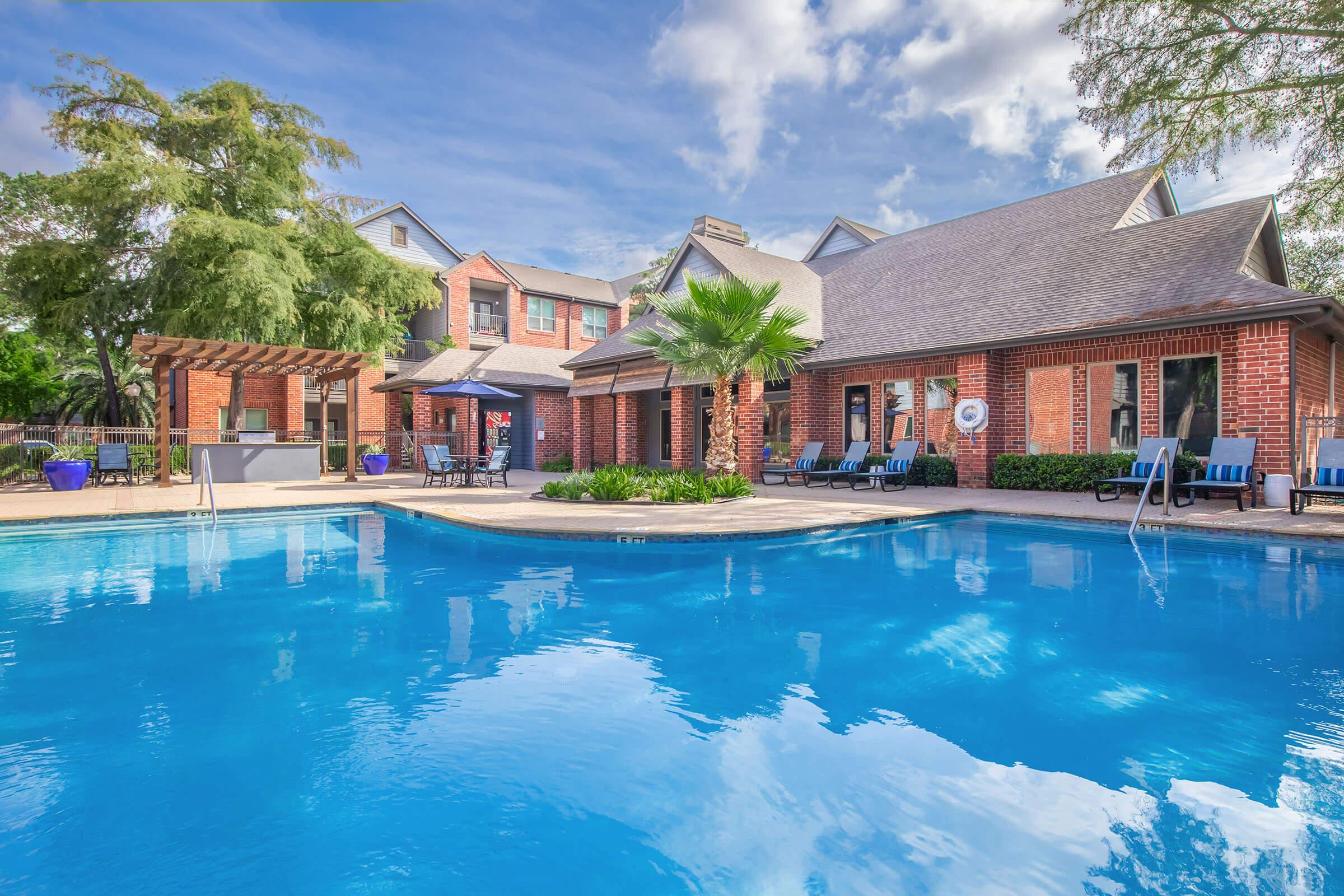 A sunny outdoor pool area featuring a clear blue pool with lounge chairs around it. In the background, there are buildings with brick facades and a covered patio. Lush greenery and palm trees add to the inviting atmosphere. The sky is partly cloudy, enhancing the bright ambiance.