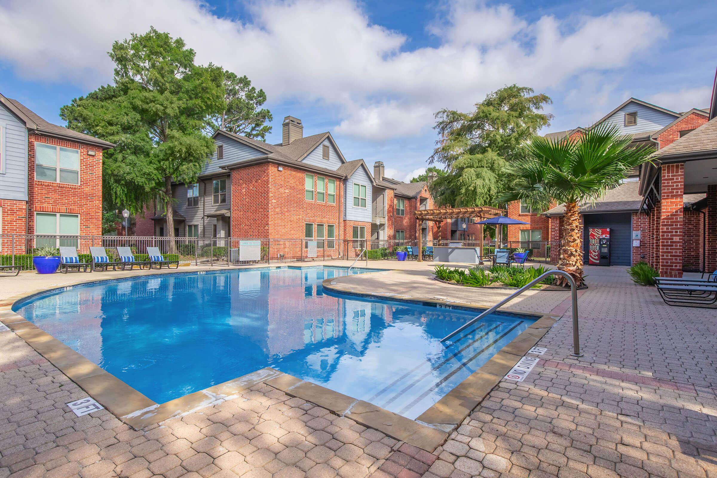 Relaxing pool area surrounded by brick apartment buildings. The clear blue water reflects the sky and surrounding trees. Lounge chairs line the poolside, and a small palm tree adds a tropical touch. A covered seating area and vending machines are visible in the background, creating a peaceful outdoor space.