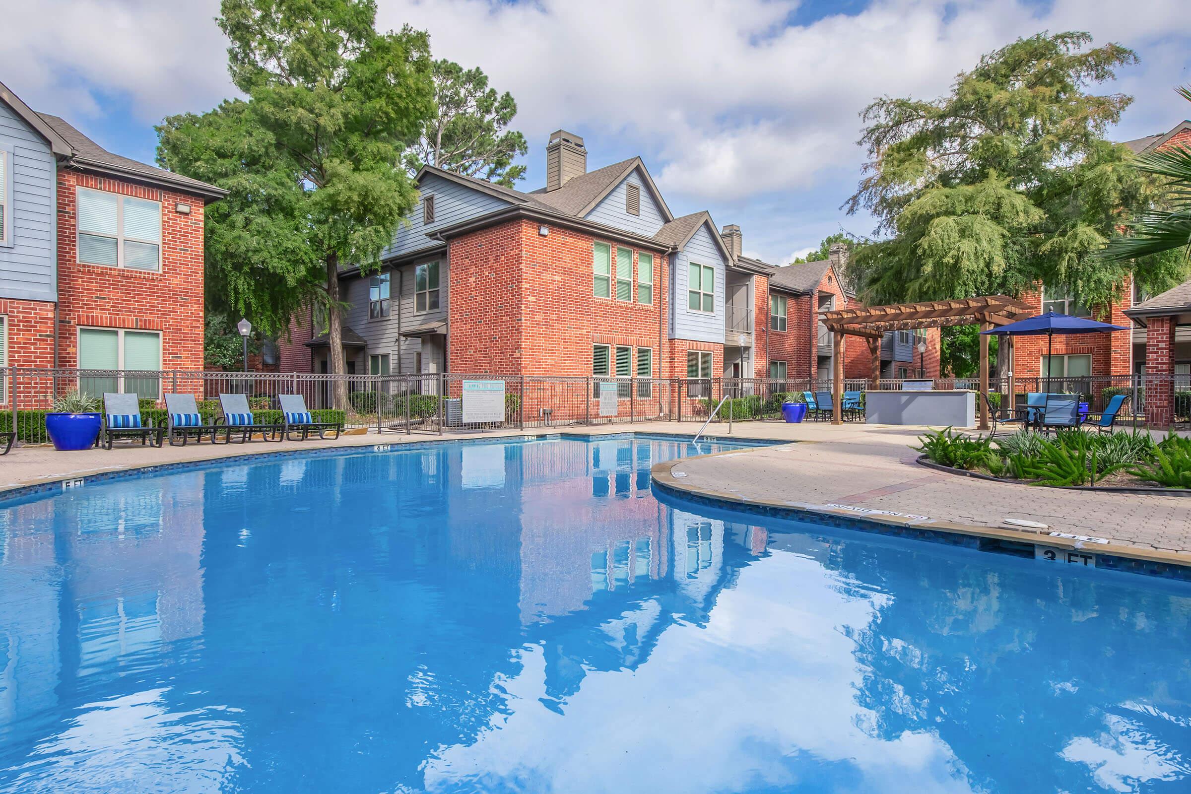 A tranquil pool area surrounded by brick apartment buildings, lush greenery, and lounge chairs. The water reflects the clear sky and nearby trees, creating a serene atmosphere. Outdoor seating and a pergola enhance the inviting outdoor space.
