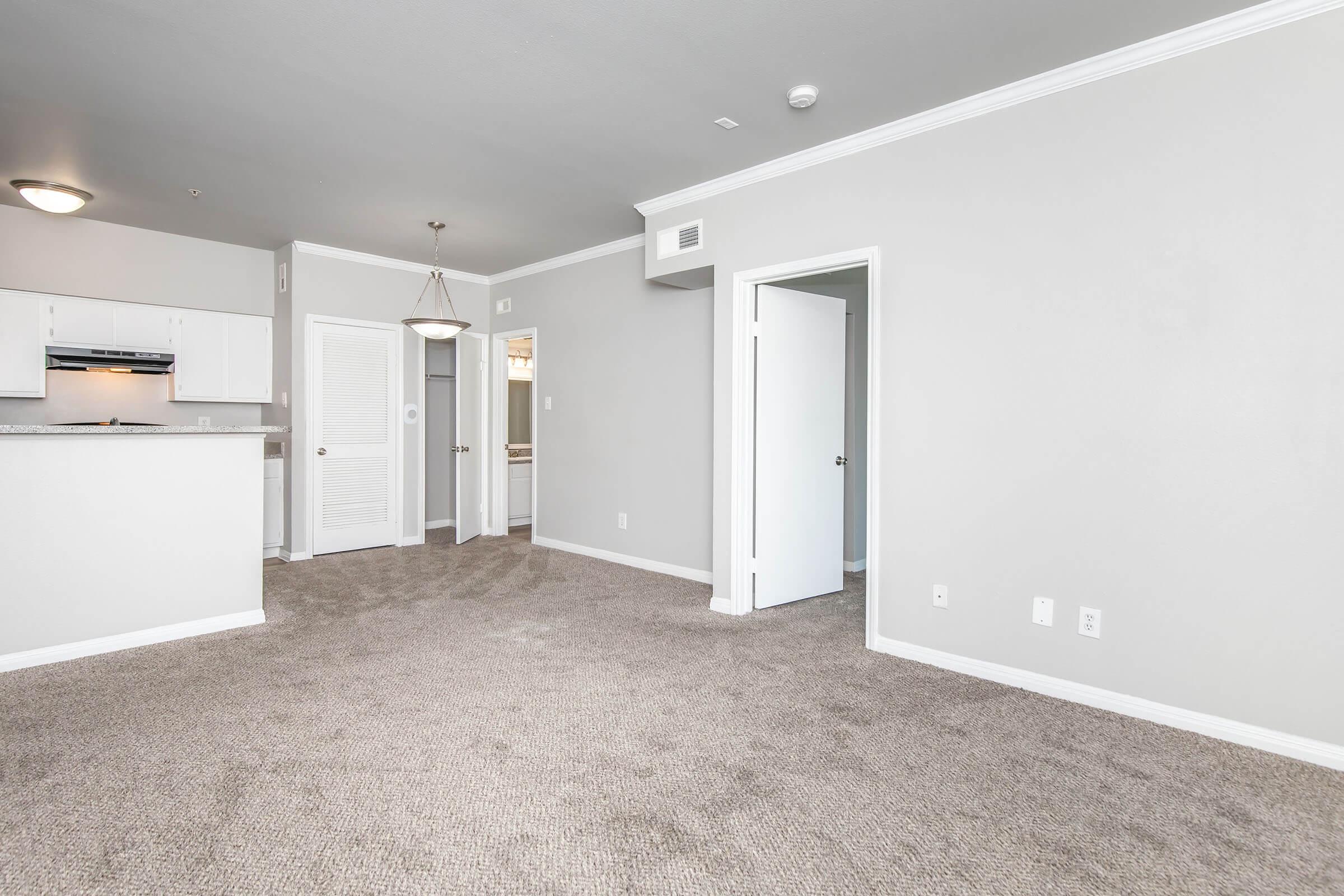 Spacious living area with light gray walls and carpet flooring. A kitchen area is partially visible on the left, featuring a bar counter. There is a light fixture hanging above the entryway, and doorways lead to another room and a bathroom. The space appears bright and inviting, suitable for modern living.