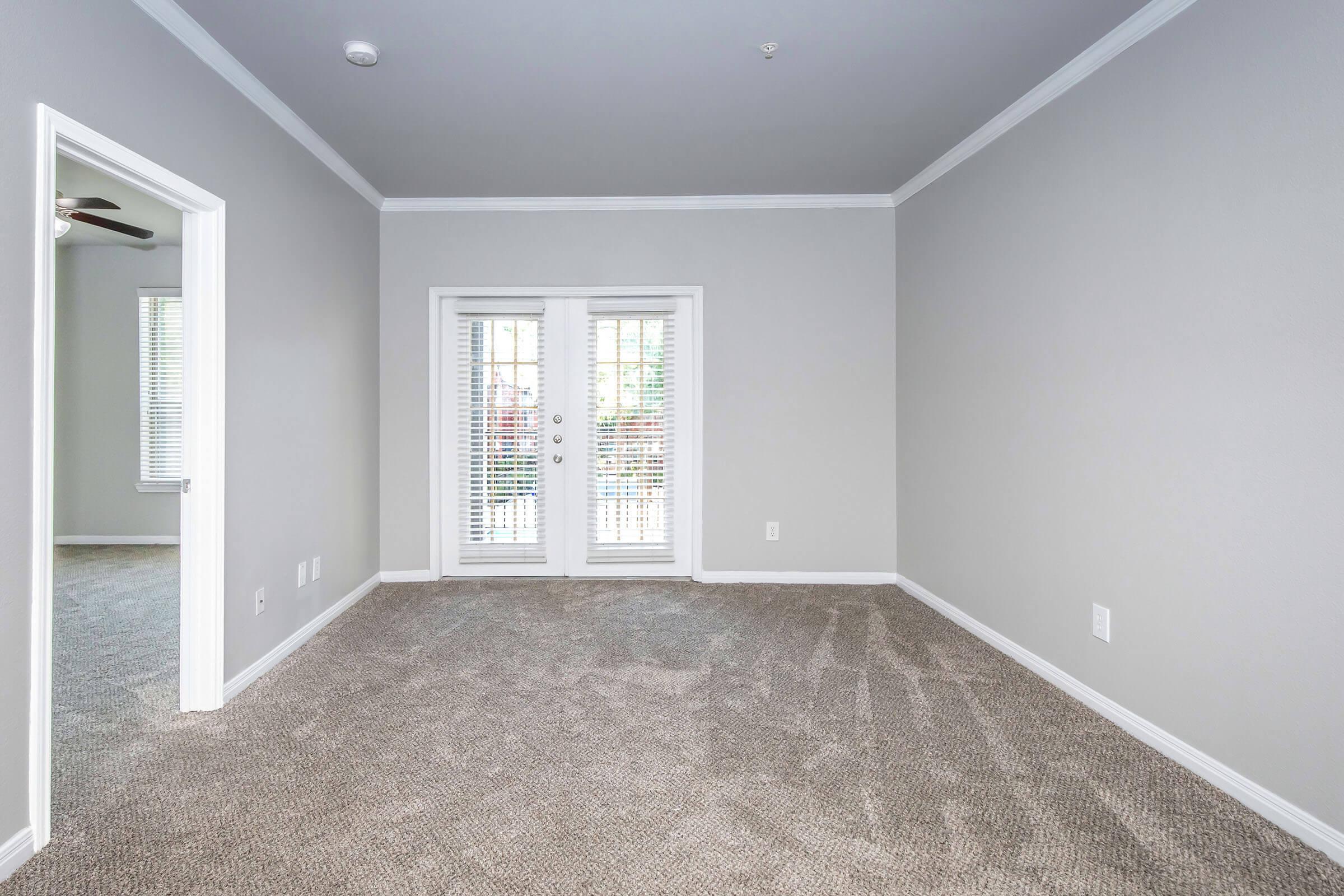 Empty interior room with light gray walls and beige carpet. There are double doors with glass panes that lead to an outdoor area, allowing natural light in. A ceiling fan is visible, and another room can be seen through an open doorway. Overall, the space appears clean and well-maintained.