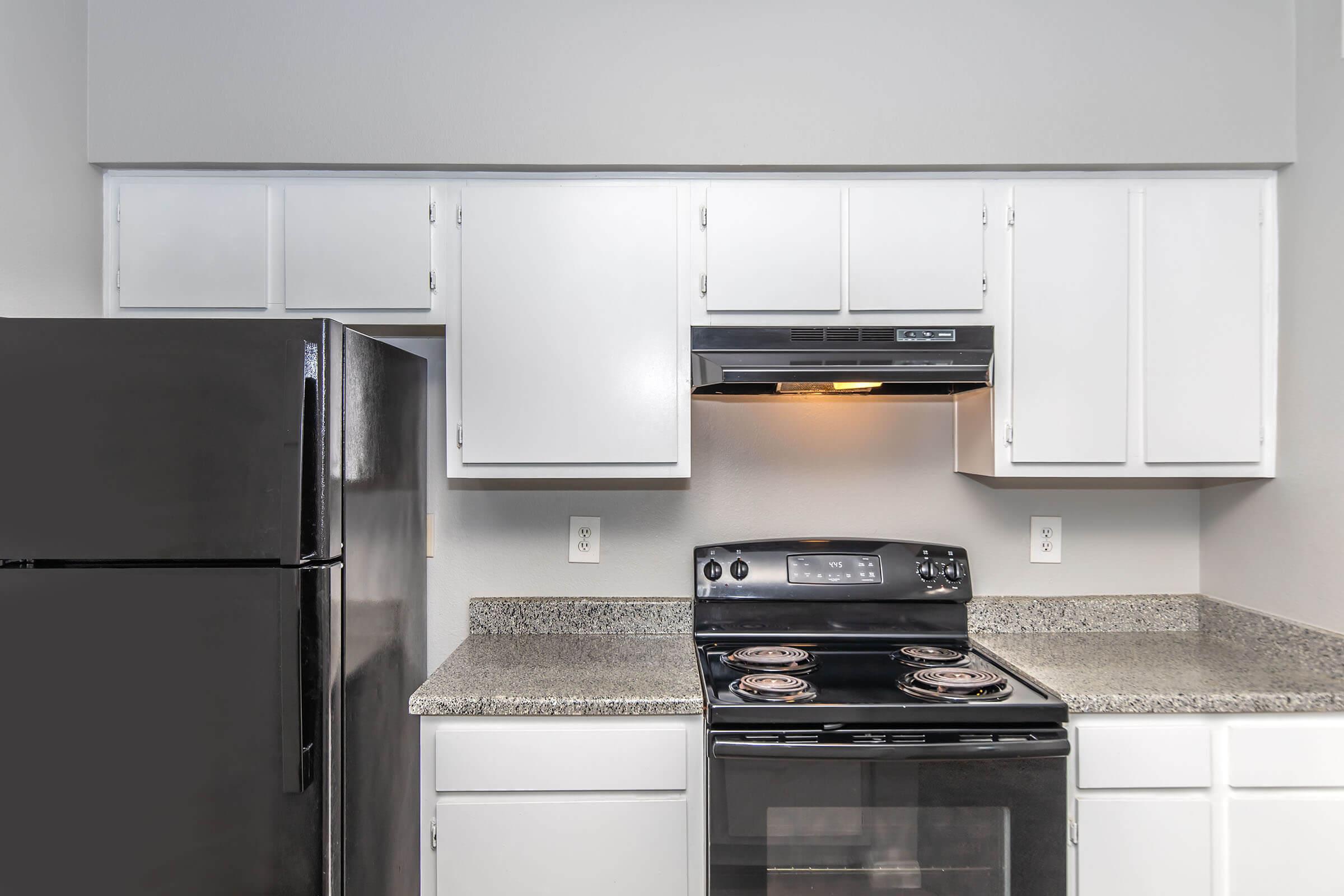 A modern kitchen featuring a black refrigerator, a black stove with an oven, and white cabinets above a granite countertop. The kitchen is well-lit with an overhead light and has a clean, minimalist design.