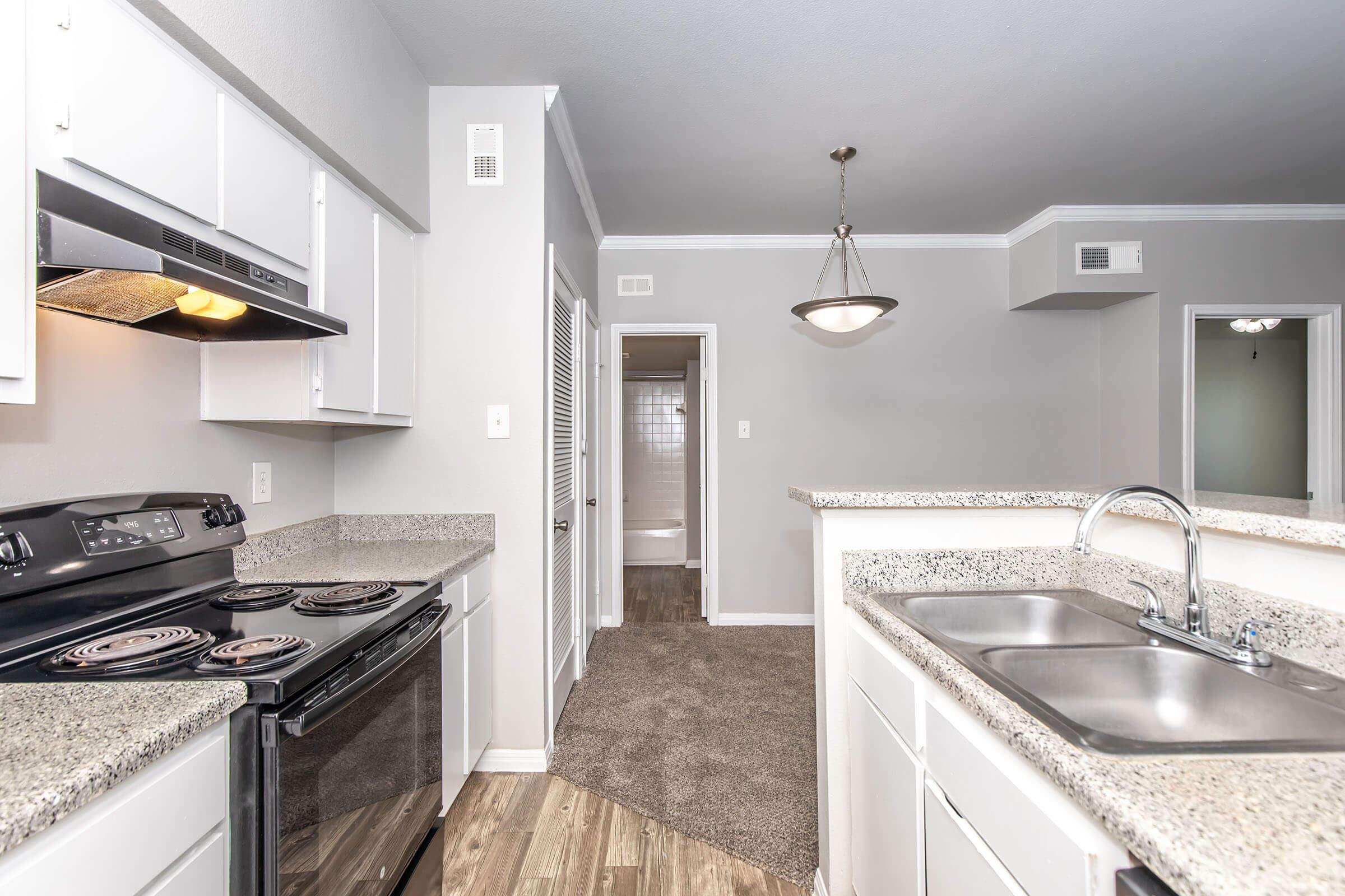 Modern kitchen with granite countertops and a black stove, featuring stainless steel appliances. There's a double sink with a faucet, and a hallway leading to another room. The flooring is a mix of wood and carpet, with neutral-colored walls and bright overhead lighting.