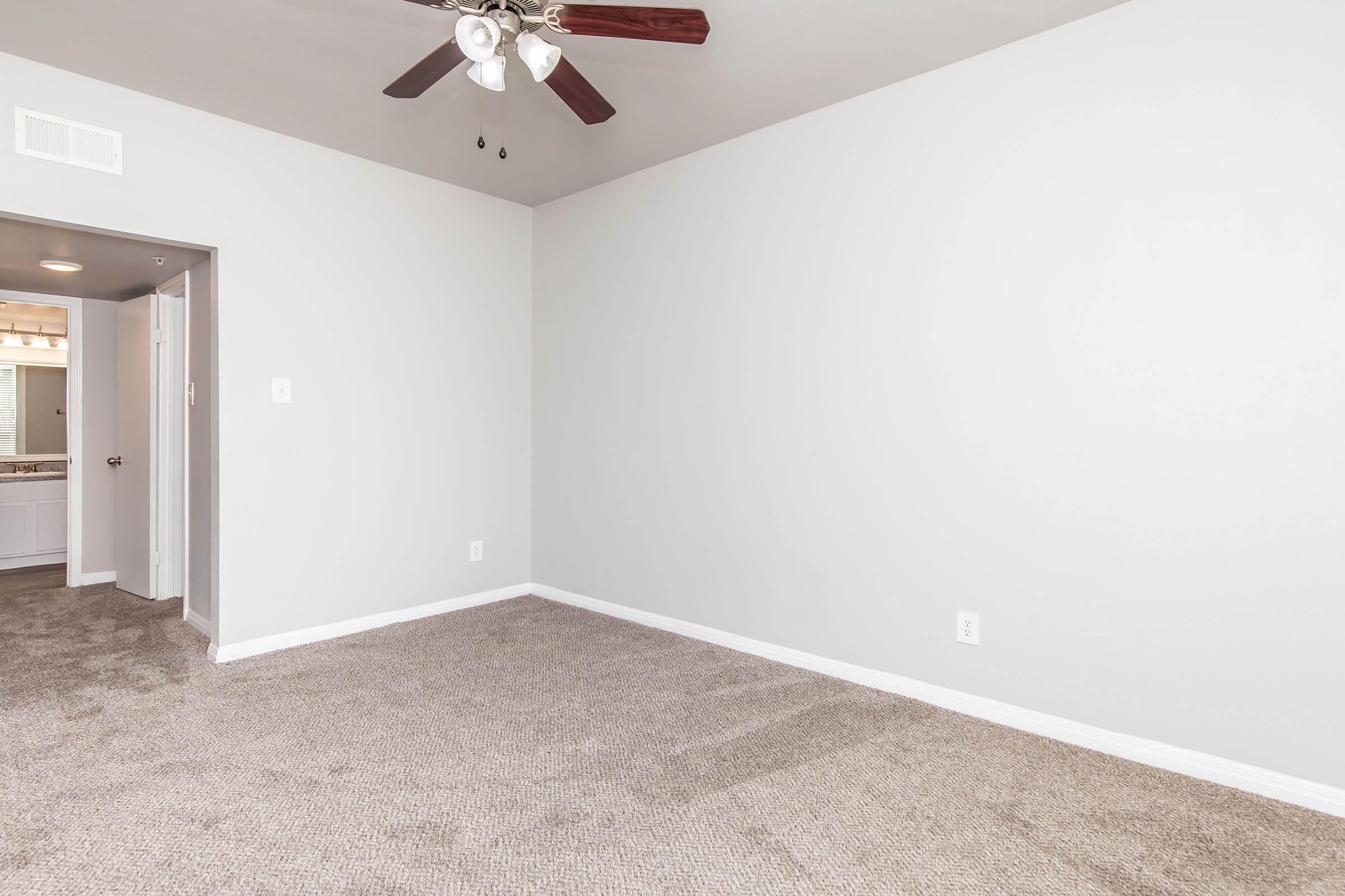 A spacious, empty room featuring light gray walls and a ceiling fan with three blades. The floor is covered in beige carpet. A doorway leads to another area, potentially a bathroom or kitchen. Natural light brightens the space, creating an airy feel. The room is ideal for furnishing and personalizing.