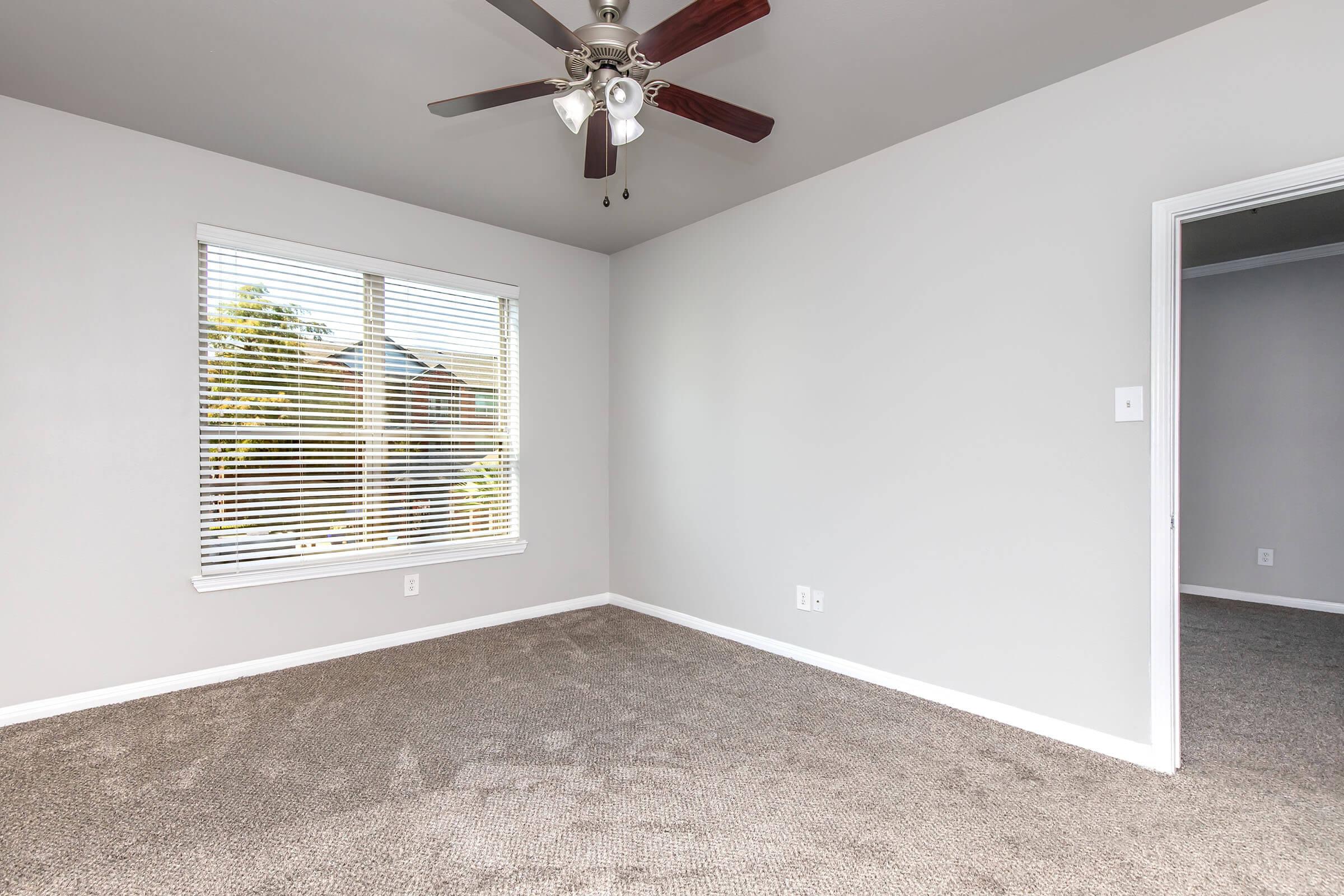 A bright, empty room with light gray walls and a ceiling fan. It features a window with white blinds, allowing natural light to enter, and a plush carpet. A doorway on the right leads to another room, suggesting an open and spacious layout.