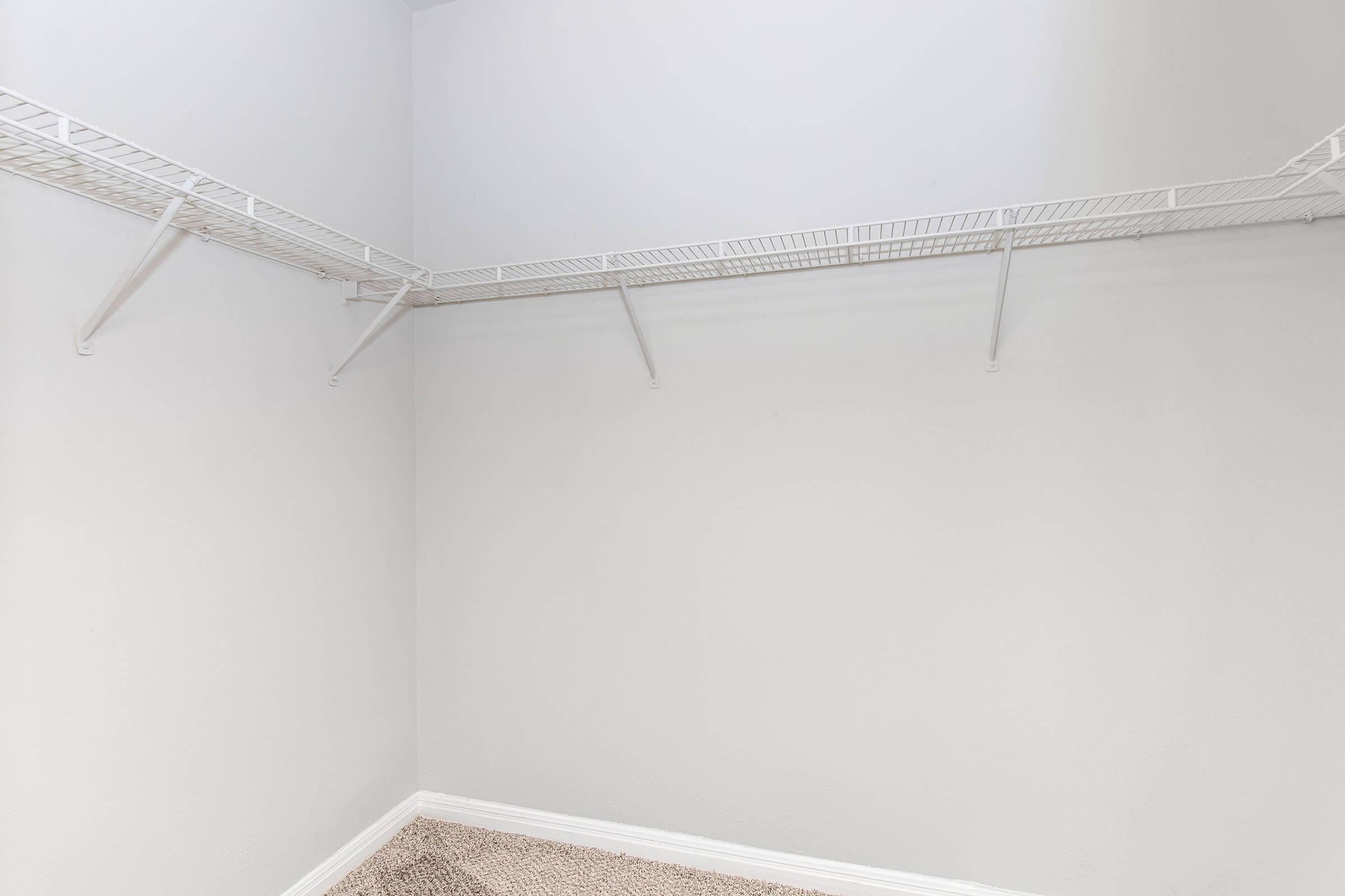 A spacious, empty closet with white walls and a carpeted floor. The closet features white wire shelving running along two walls, providing ample storage space, but currently has no items placed on the shelves. The overall look is clean and uncluttered.