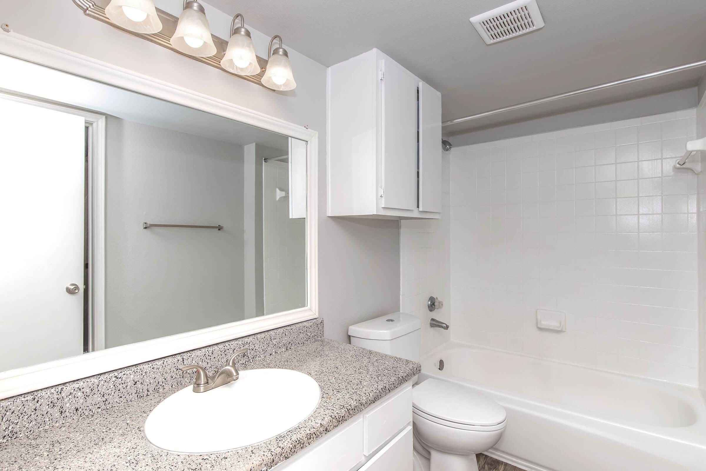 A clean, modern bathroom featuring a white bathtub and shower combo, a toilet, and a countertop with a sink. The walls are light-colored, and there is a large mirror above the sink with ambient lighting. A cabinet is visible above the toilet, and there is a towel bar on the wall.