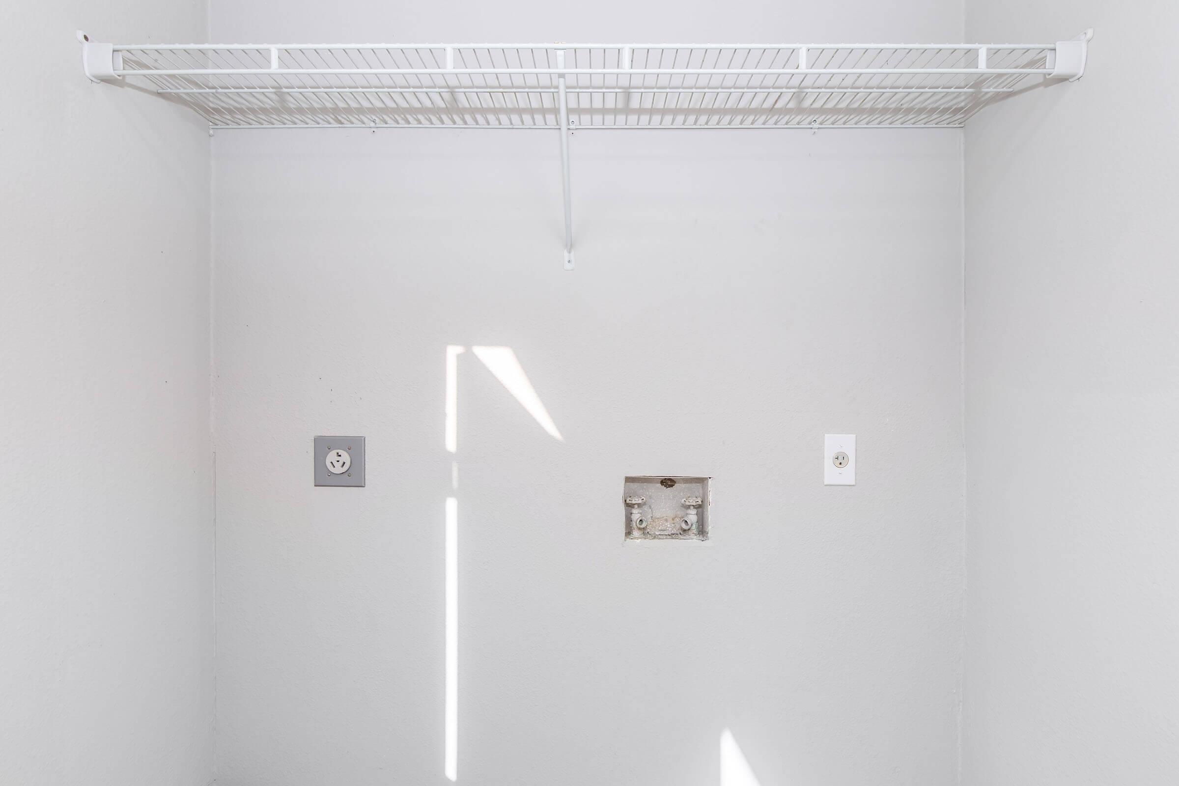 A small, empty closet with a white wire shelf mounted on the wall. The walls are painted in a light color, with a power outlet on the left and a blank electrical box in the center. A beam of sunlight casts a shadow across the wall, creating a bright, minimalistic atmosphere.