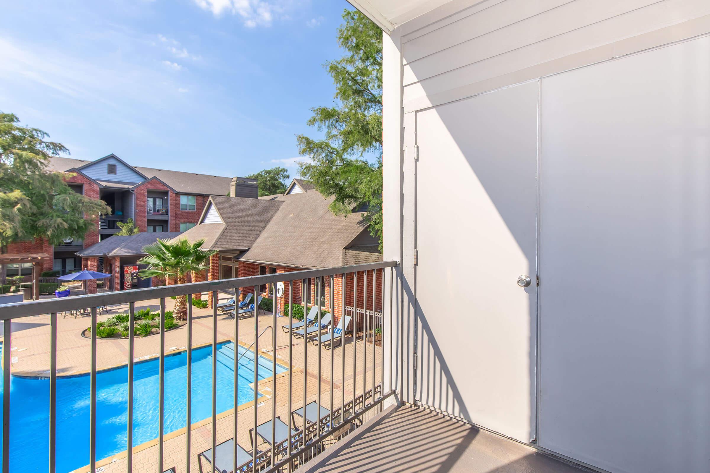 View from a balcony overlooking a pool area surrounded by residential buildings. The pool is blue with lounge chairs nearby, and there are trees in the background under a clear sky. The scene is bright and inviting, suggesting a pleasant outdoor atmosphere.
