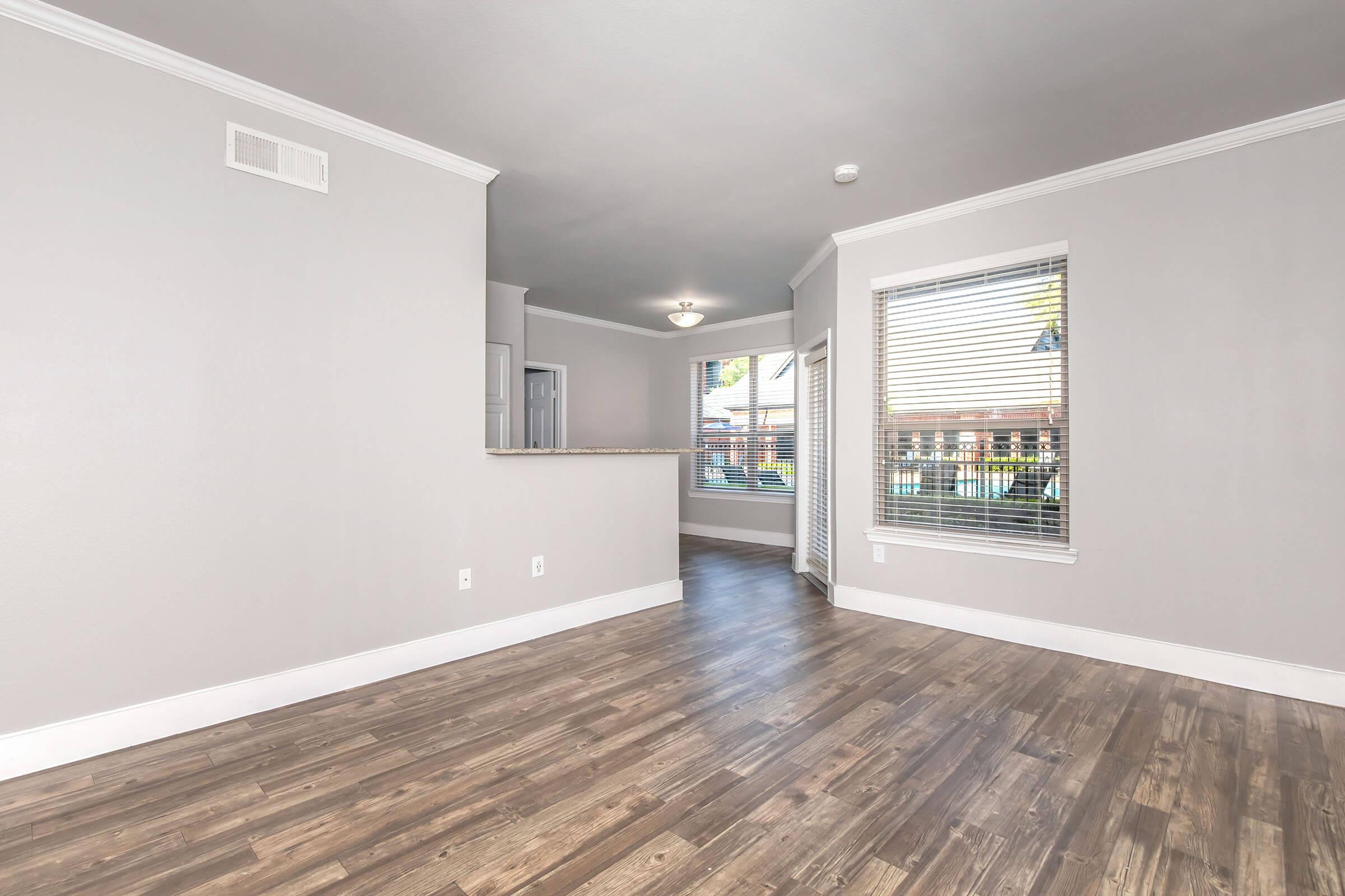 Spacious, modern living area featuring light gray walls and wood-like flooring. A large window allows natural light to flood the room, with views of the outdoors. The open layout includes a kitchen nook visible in the background, enhancing the inviting atmosphere. Ideal for relaxed living and entertaining.