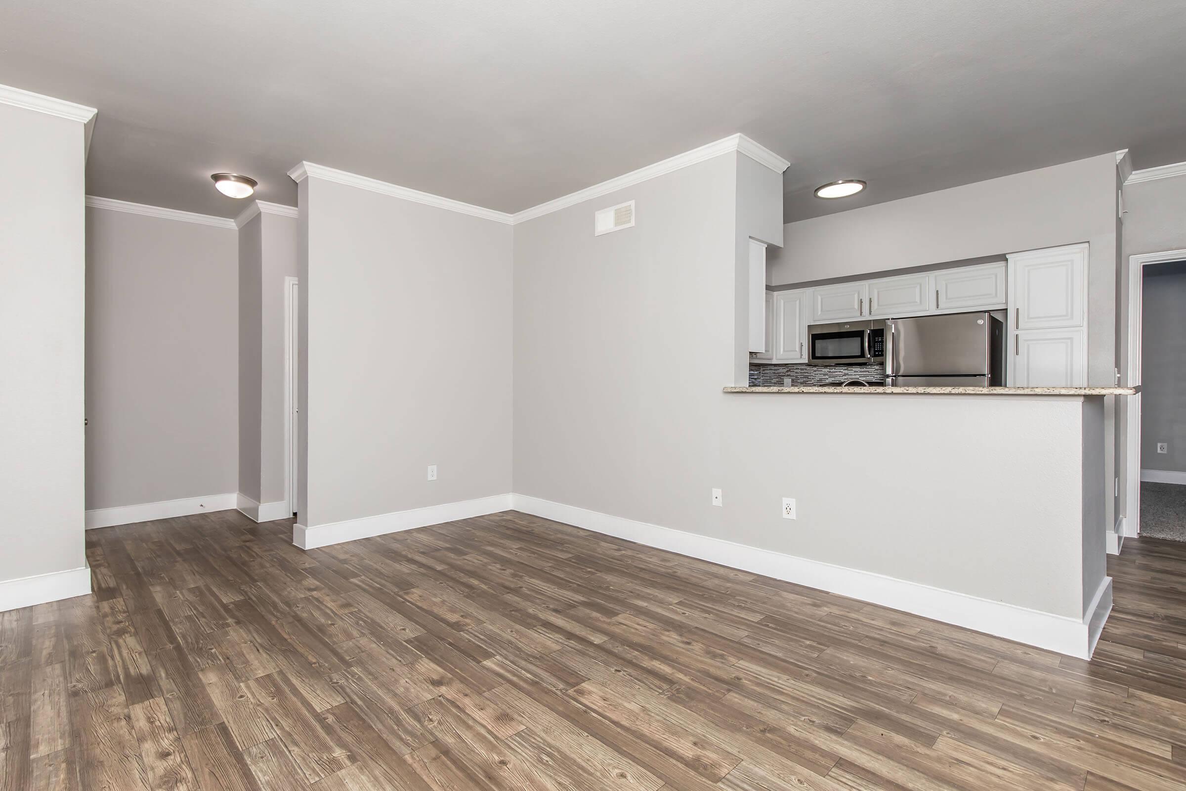 A spacious empty living area featuring light gray walls, hardwood floors, and crown molding. The open layout includes a kitchen area with white cabinetry, a granite countertop, and a stainless steel refrigerator. Natural light fills the room, highlighting its clean and modern design.