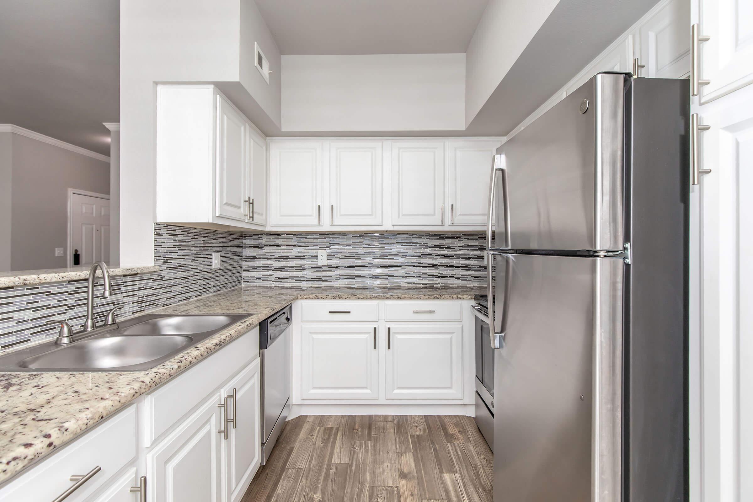 Modern kitchen featuring white cabinetry, a stainless steel refrigerator, and a double sink. The countertops are made of speckled granite, and the backsplash consists of patterned mosaic tiles. The space has a warm ambiance with wooden flooring and ample natural light.