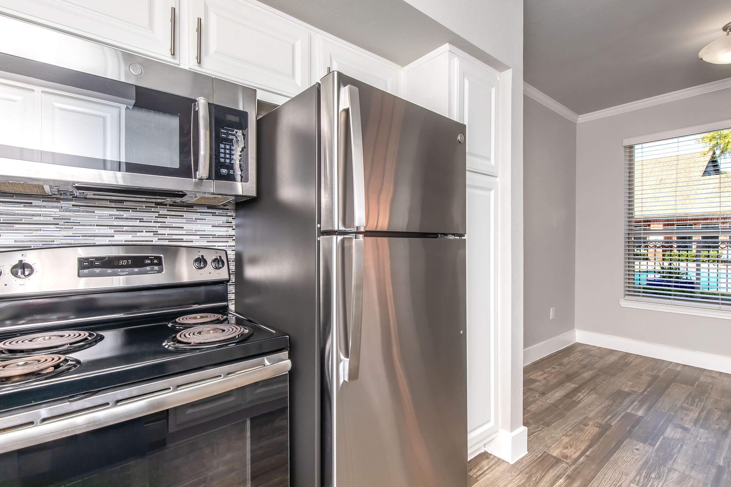 Modern kitchen corner featuring a stainless steel refrigerator next to a black stove with four burners. The walls are painted a light gray, and white cabinetry complements the design. A window with blinds allows natural light to enter, showcasing a glimpse of an outdoor area beyond.
