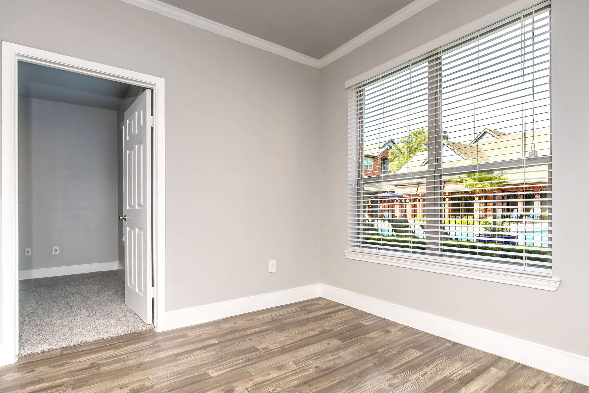 A well-lit empty room with gray walls and wood-style flooring. A large window with blinds allows natural light to enter, revealing a view of outdoor greenery. An open door leads to another space, suggesting a connected layout. The overall atmosphere is clean and inviting, ideal for personal decoration.
