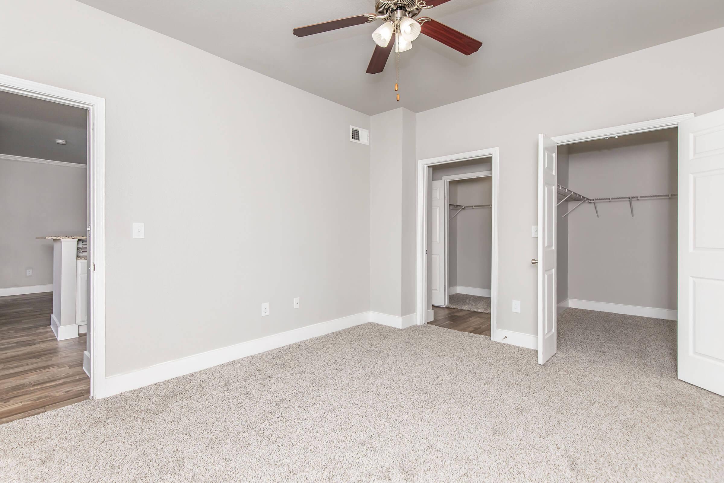 A spacious, empty bedroom featuring light gray walls, plush carpet, and a ceiling fan with wooden blades. There are two doorways: one leading to a closet with a hanging rod and the other opening to a hallway. Natural light highlights the room's inviting atmosphere.