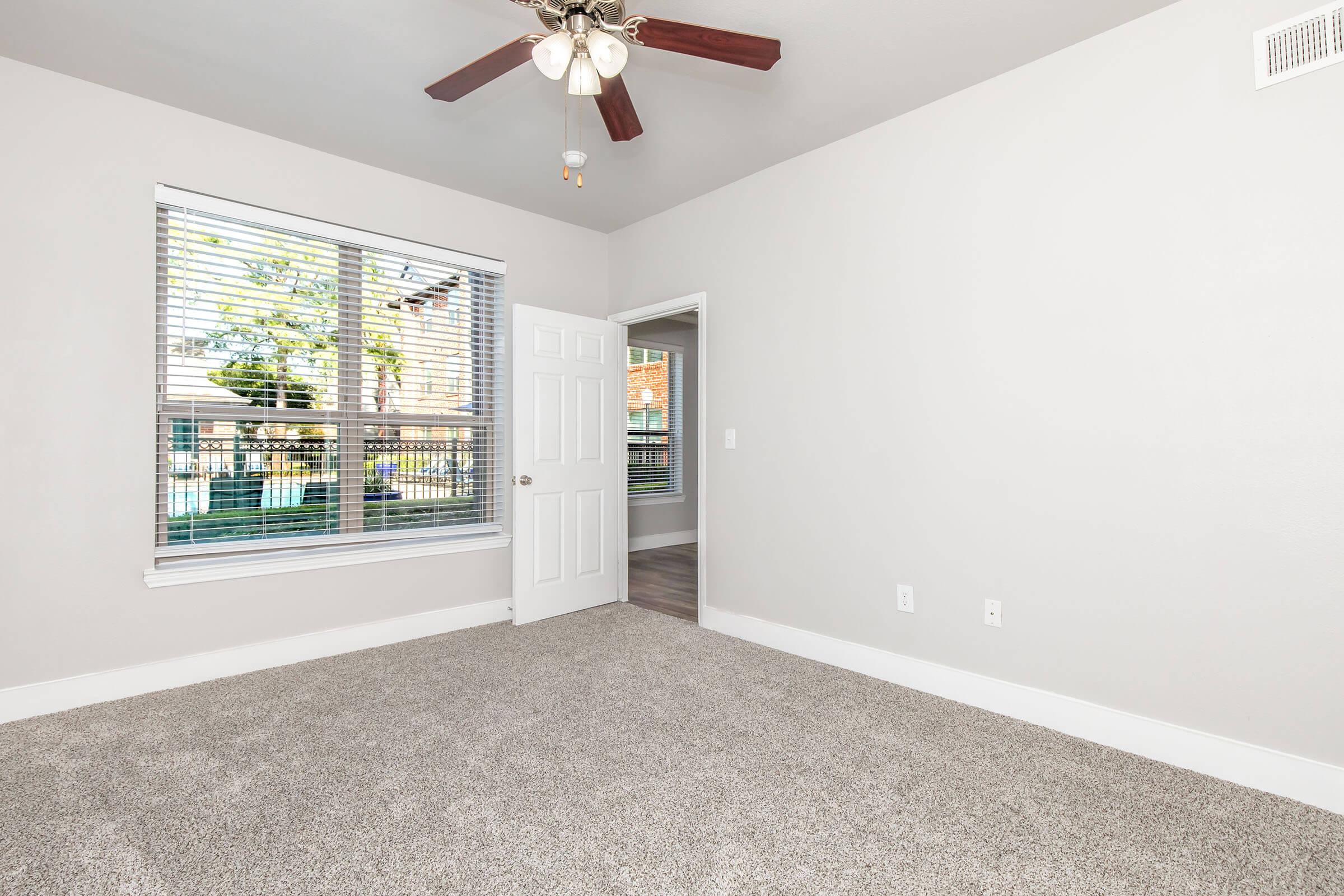 A bright and spacious room with light gray walls and carpeted flooring. A ceiling fan hangs from the ceiling, and there is a window with blinds allowing natural light. A doorway leads to another room, creating an open feel in the space.
