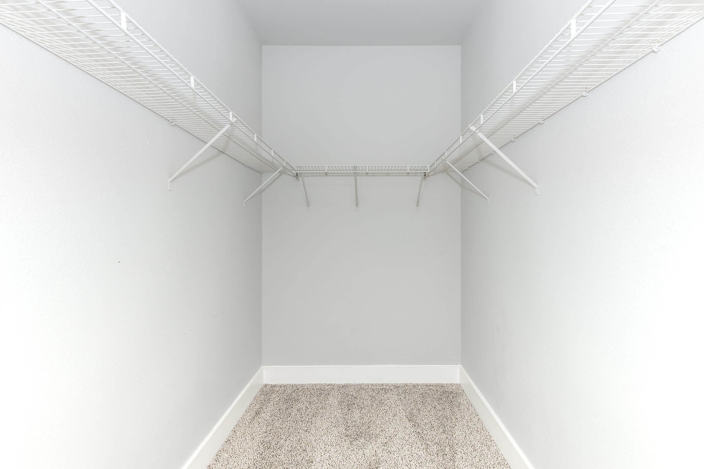 Empty closet featuring two white wire shelves along the walls. The walls are painted a light color, and the floor is covered with beige carpet. There are no items or decorations present, creating a spacious, minimalist appearance.