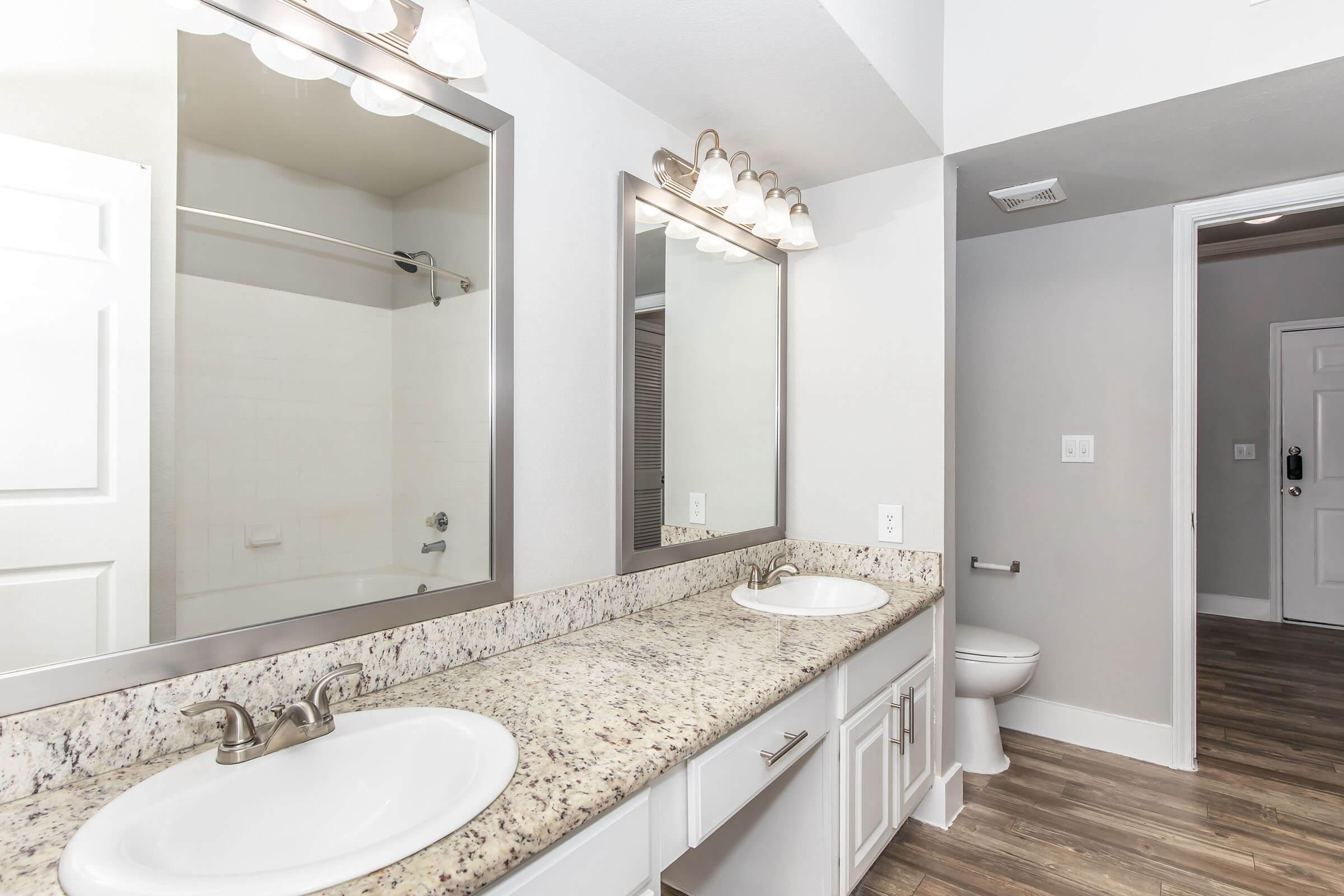 A modern bathroom featuring double sinks with a granite countertop, two mirrors, and bright overhead lighting. To the left, there is a glass shower enclosure, while the right side has a toilet. The walls are painted in a light gray, and the flooring is a wood-like laminate.