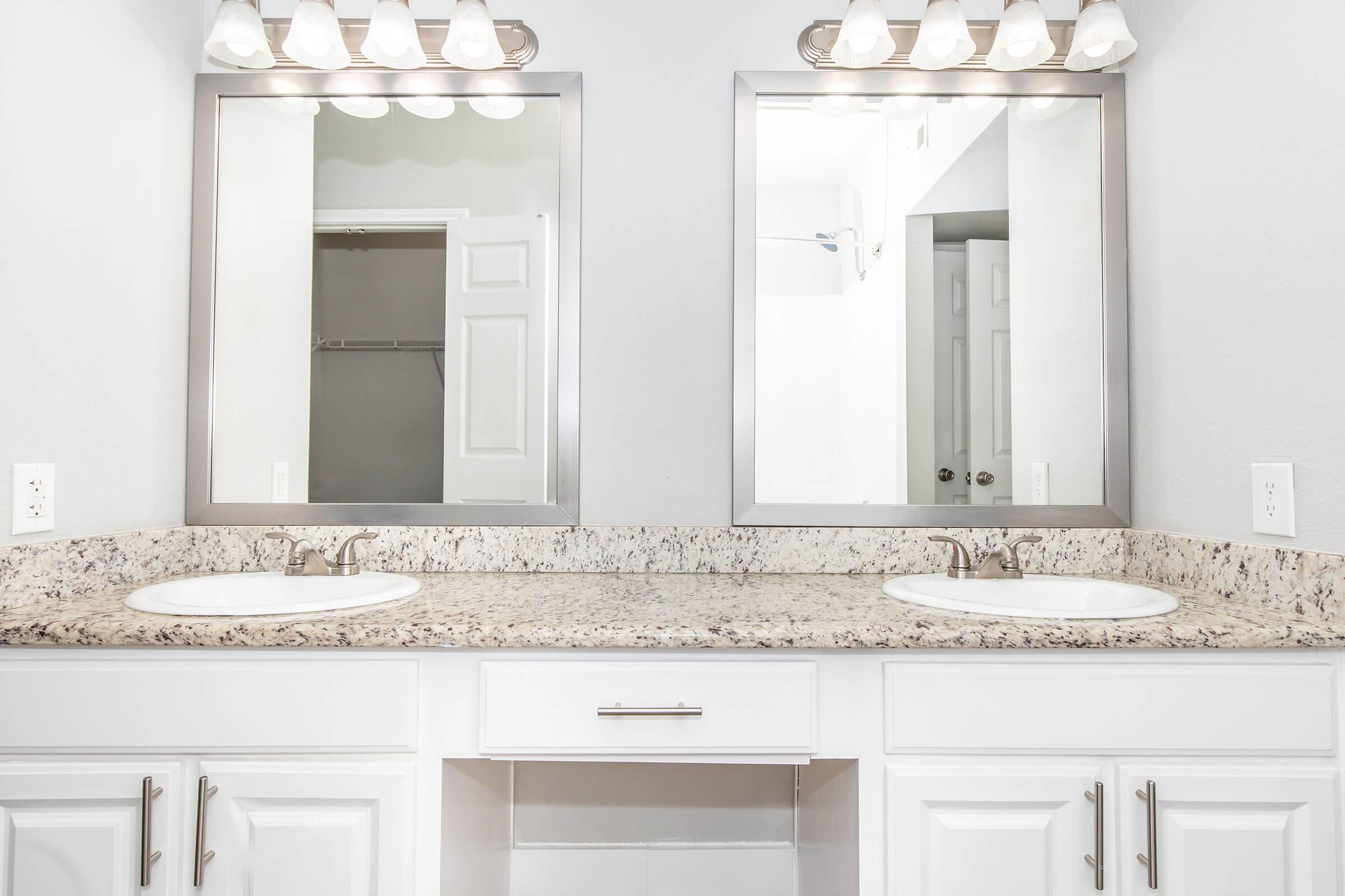 A spacious bathroom featuring a double vanity with two sinks and mirrors above. The countertops are made of speckled granite, and there's a simple white cabinet beneath. The walls are a light color, and a closet door is partially visible in the background. Natural lighting enhances the clean, modern look.