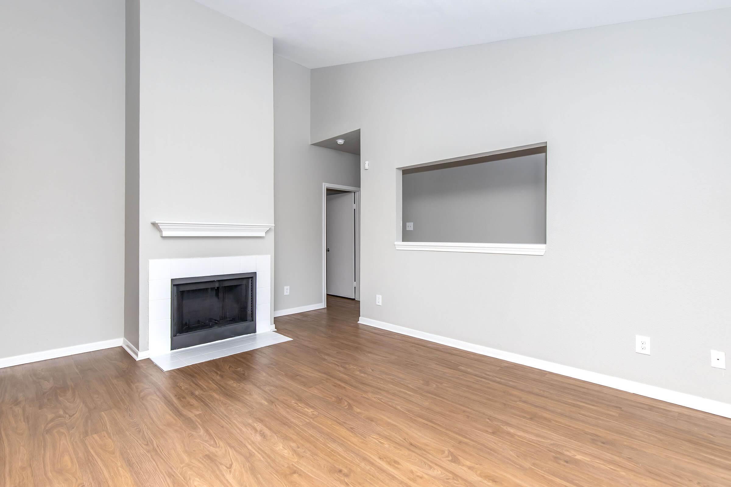 A spacious, empty living area with light gray walls and polished wooden flooring. The room features a white fireplace on the left and an opening leading to another room. Natural light illuminates the space, highlighting its airy and modern feel.