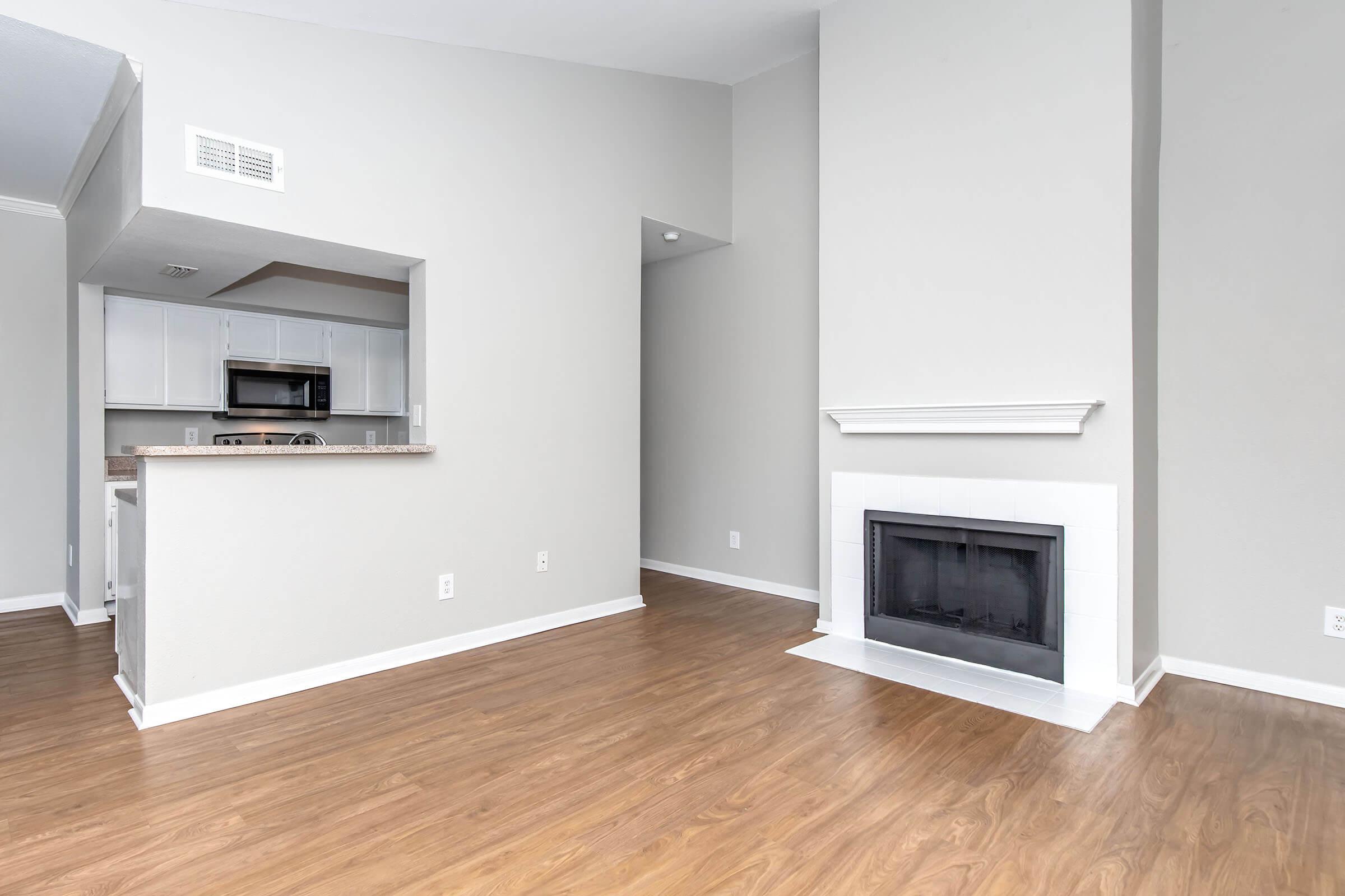 A spacious living room with light-colored walls and hardwood flooring. There is a modern fireplace with a white mantel on one side, and an open kitchen area visible in the background with white cabinets and appliances. Natural light fills the room.