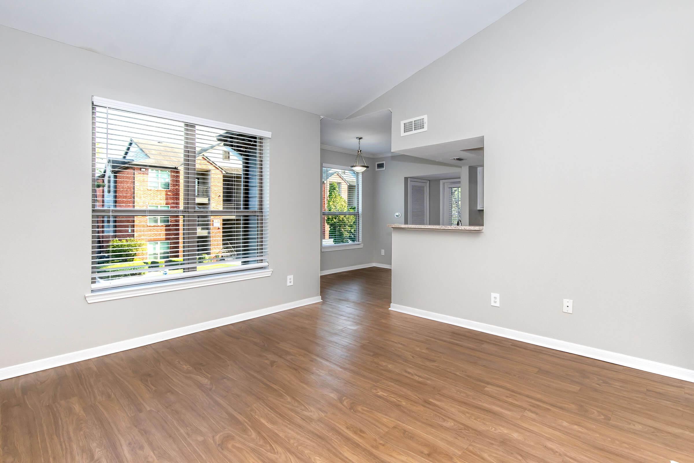 A bright, empty living room with light gray walls and hardwood flooring. A large window with white blinds lets in natural light, showcasing an exterior view of nearby buildings. The adjoining area includes an open kitchen space visible in the background, enhancing the room's airy feel.