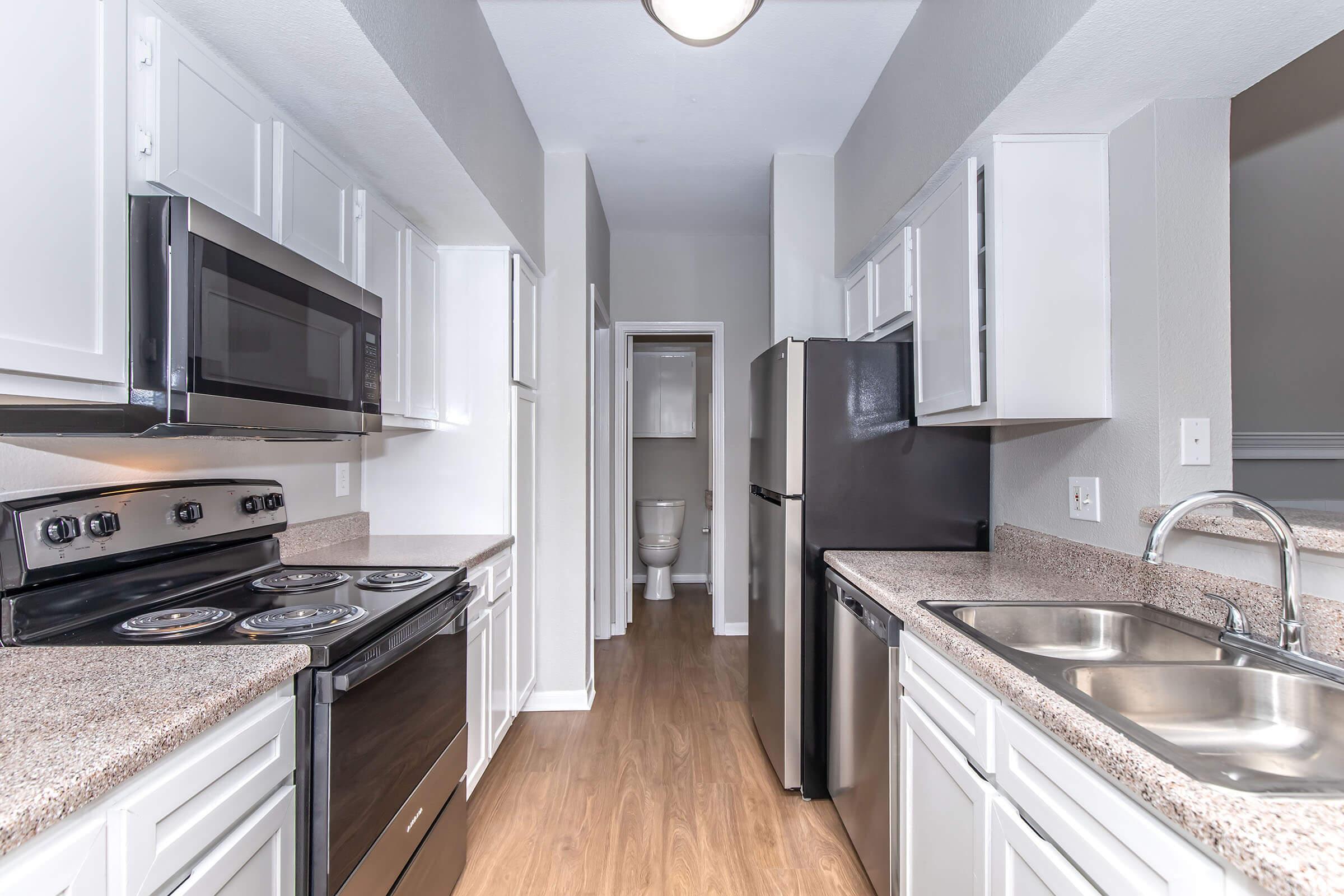 A modern kitchen featuring white cabinets, a stainless steel microwave, a black oven and refrigerator, and a granite countertop. The layout is narrow, with a doorway leading to a bathroom at the end. The flooring is light wood, creating a clean and bright atmosphere.