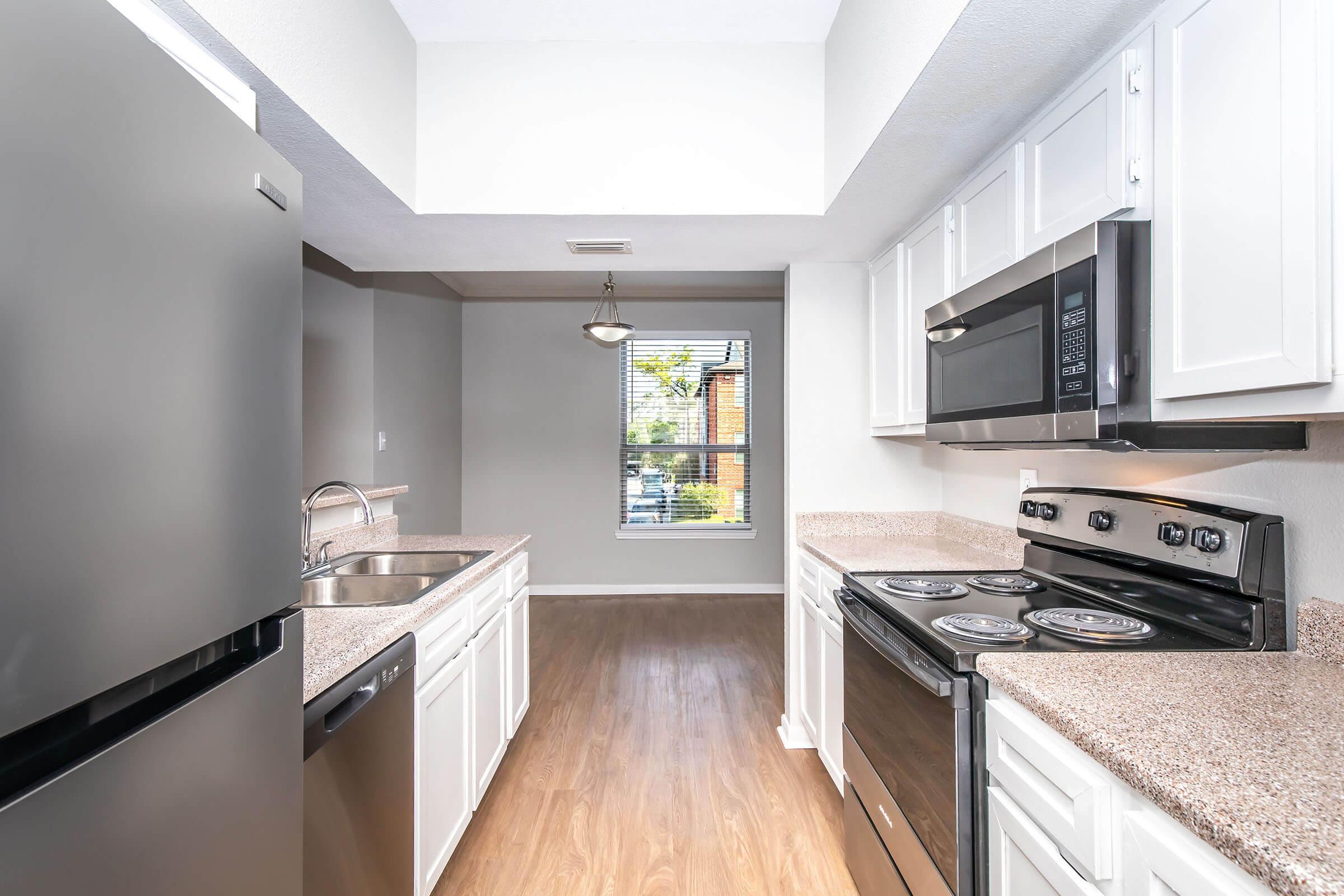 Modern kitchen featuring black and stainless steel appliances, including a refrigerator, stove, and microwave. The space has white cabinetry, granite countertops, and wood-style flooring. A window allows natural light, providing a view of the outdoors. The kitchen is well-lit with a ceiling light fixture.