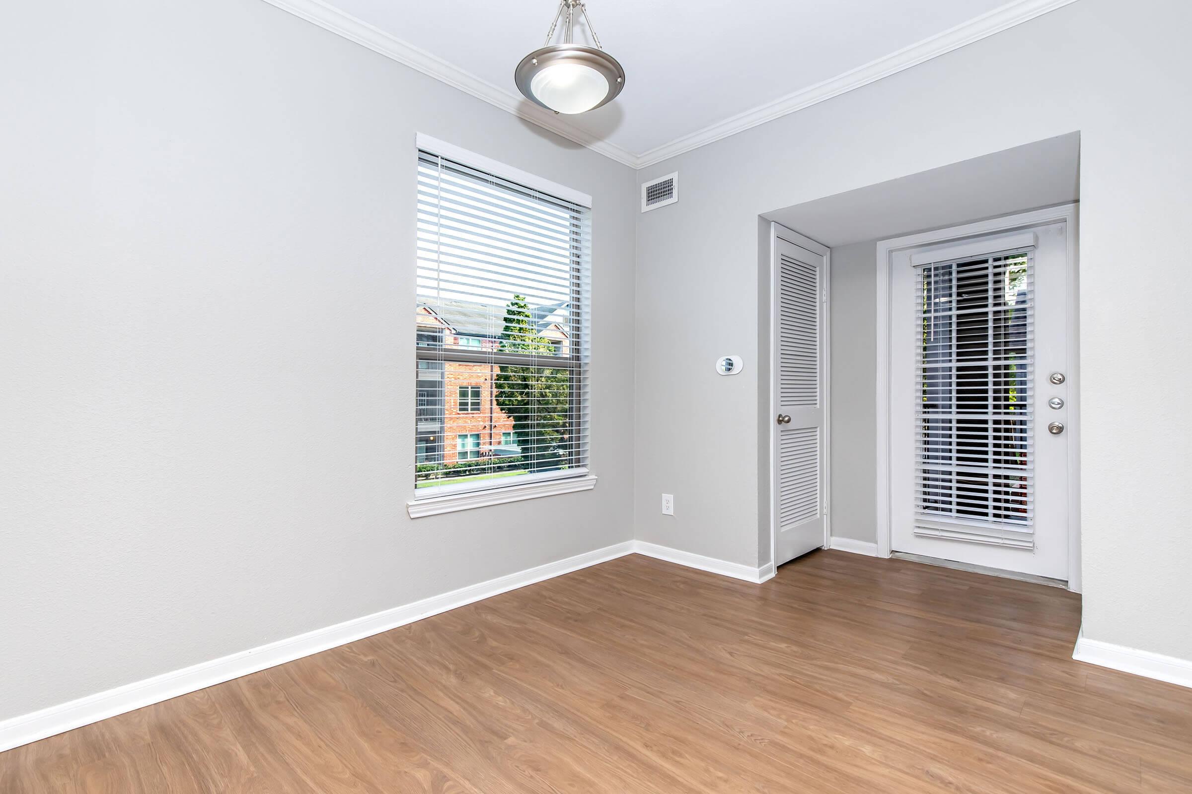 A bright, empty room with light gray walls and a wood floor. It features a window with blinds letting in natural light, a door leading outside, and a closet door. A ceiling light hangs above, illuminating the space. The overall ambiance is clean and modern, ideal for various uses.