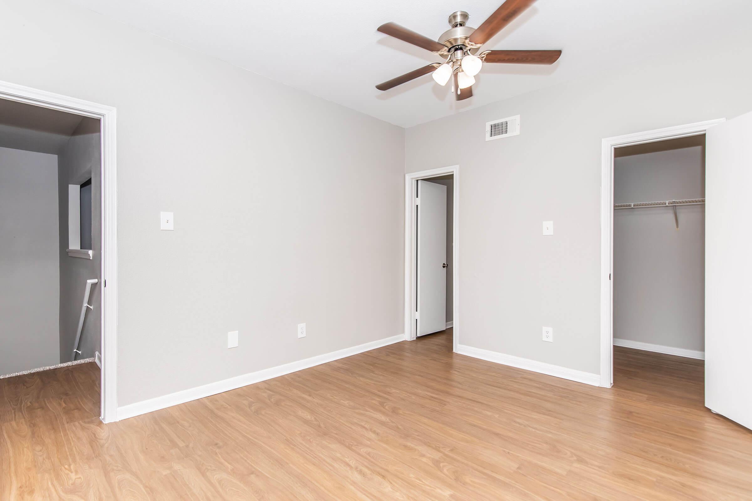 Empty room featuring light-colored walls and wooden flooring. A ceiling fan with wooden blades hangs from the ceiling. Two doorways lead to adjacent spaces, one with a closet. The room is well-lit and casual, making it suitable for various uses such as a living area or bedroom.