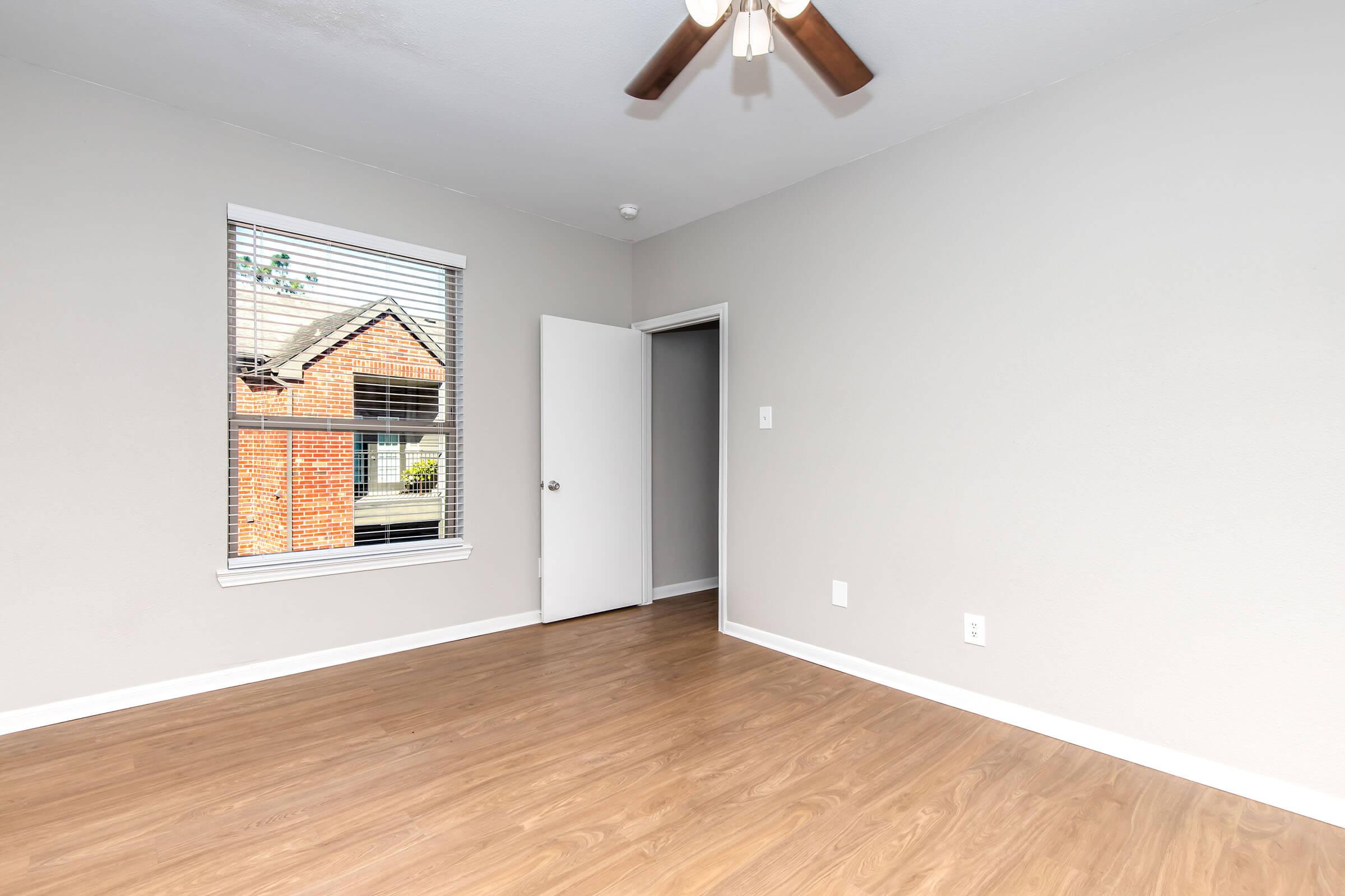 Empty room with light-colored walls and wooden flooring. A window with blinds lets in natural light, showing a glimpse of a brick exterior outside. There is a ceiling fan and an open door leading to another space. The room appears clean and well-lit, creating a spacious feel.
