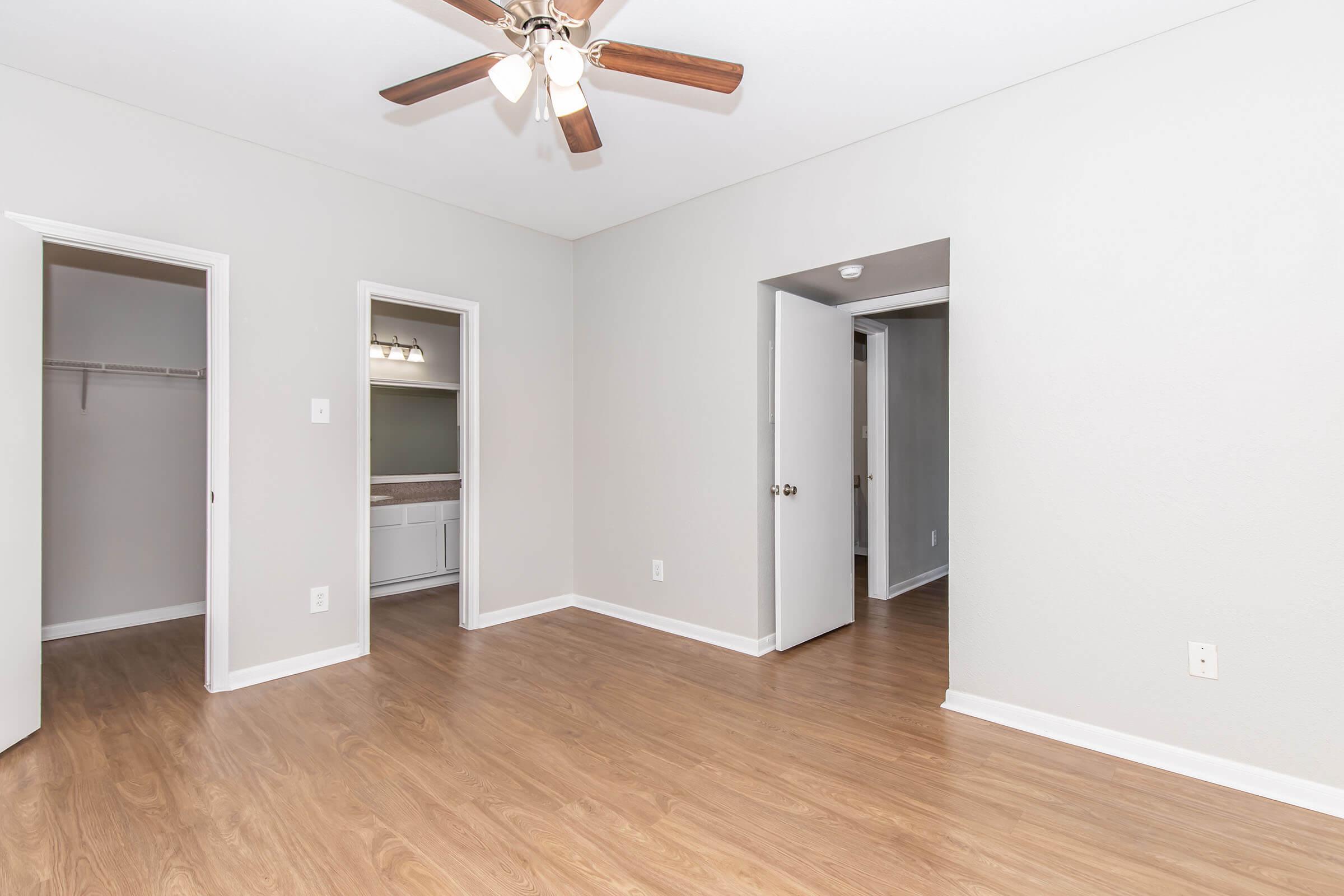 A spacious, empty room with light gray walls and a ceiling fan. The floor is a light wood laminate. There are two closed doors leading to adjacent rooms, and a third open doorway leading to a bathroom area visible in the background. The room is well-lit and has a clean, modern feel.