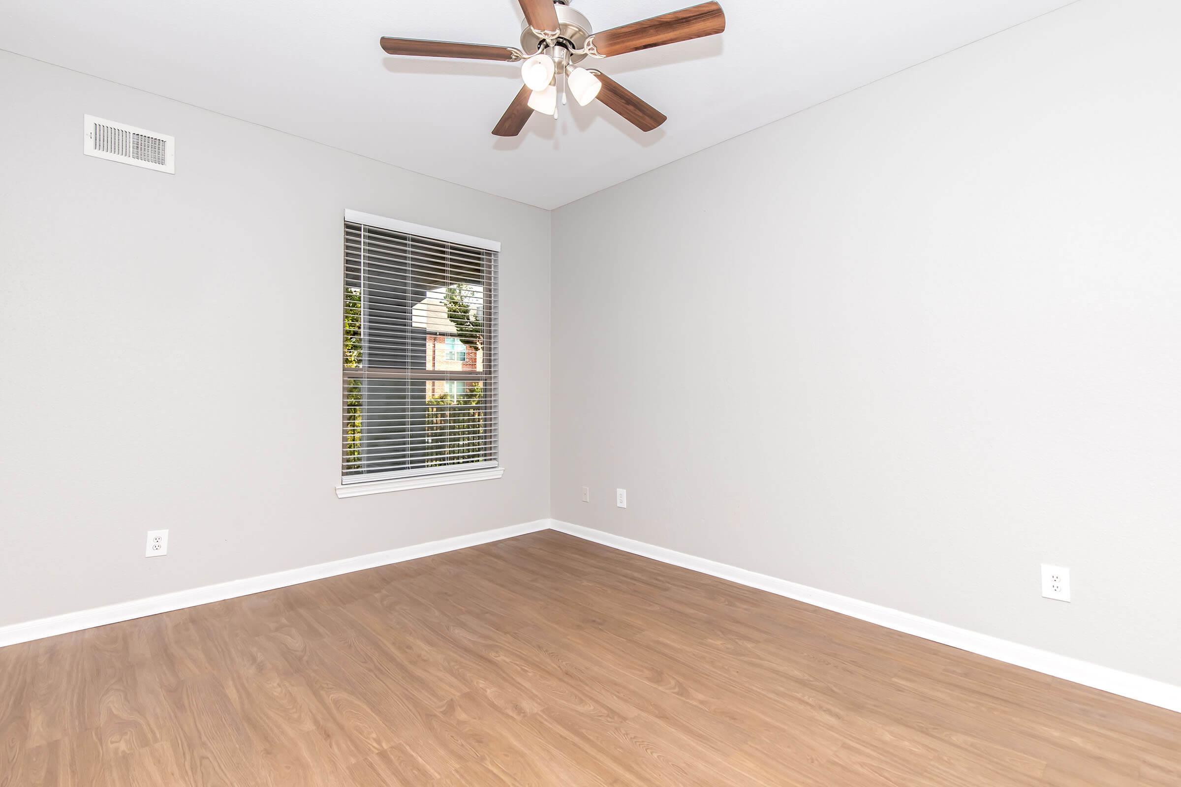 A spacious, empty room with light gray walls and a wooden floor. A ceiling fan with five blades is mounted above, and a large window with blinds lets in natural light. The room has a simple, modern design with no furniture.