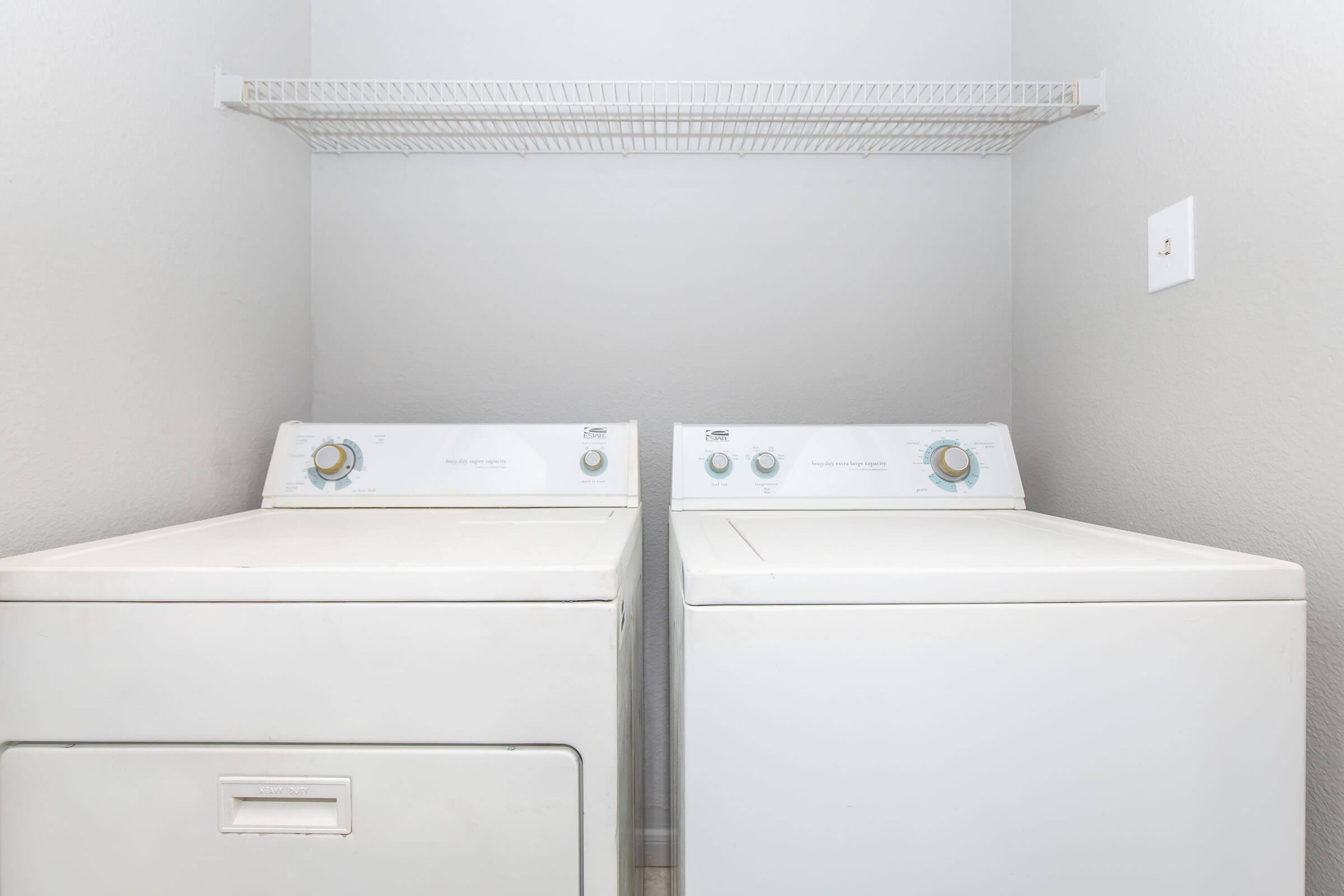 A bright laundry room featuring a white washer and dryer set side by side, with a gray wall in the background. Above the appliances is a metal wire shelf for storage. The overall space is clean and organized, highlighting a practical and functional laundry area.