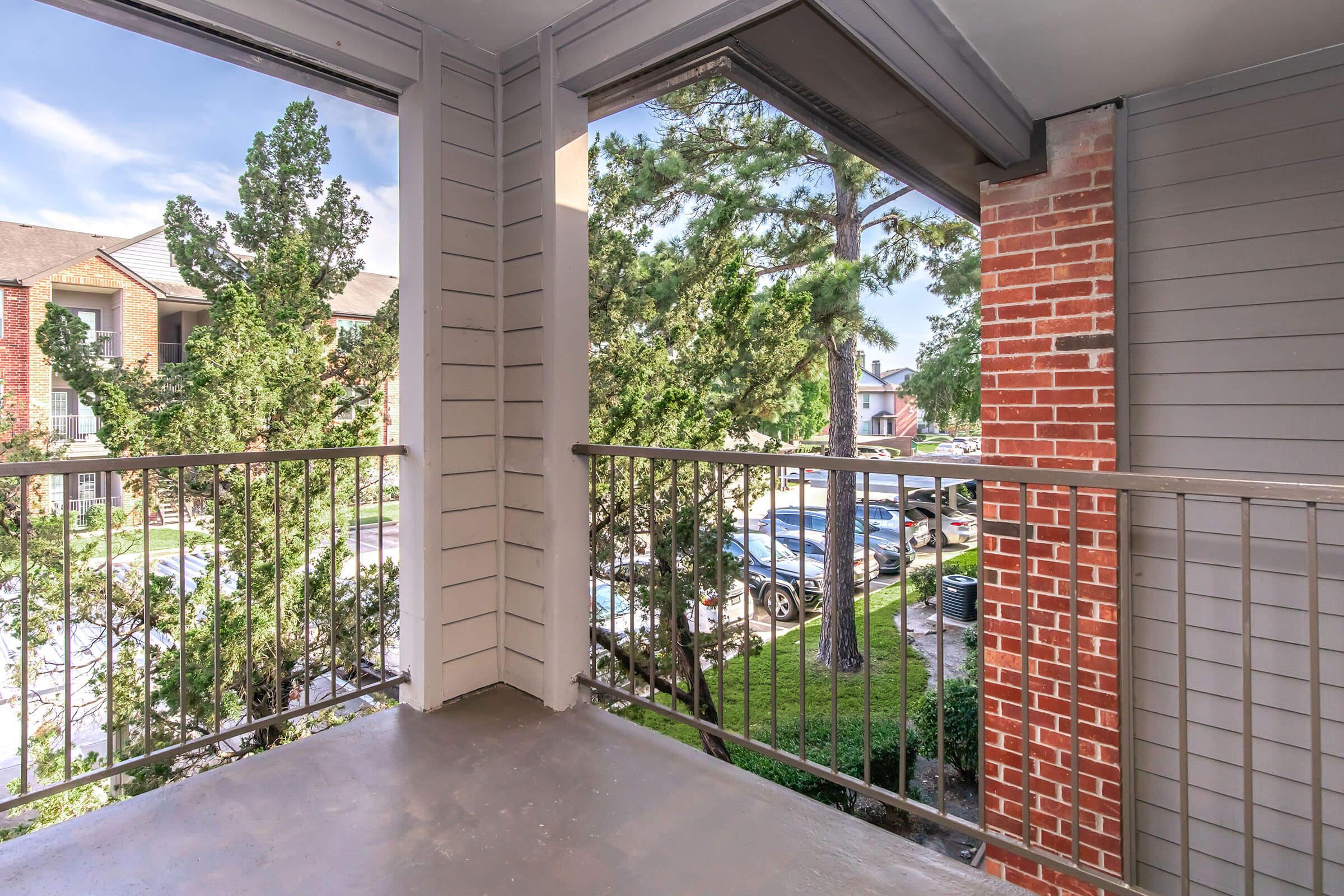 View from a balcony overlooking a green area with trees and parked cars. The balcony has a metal railing and is part of a multi-story building with brick and siding exterior. The sky is clear and blue, suggesting a bright day.