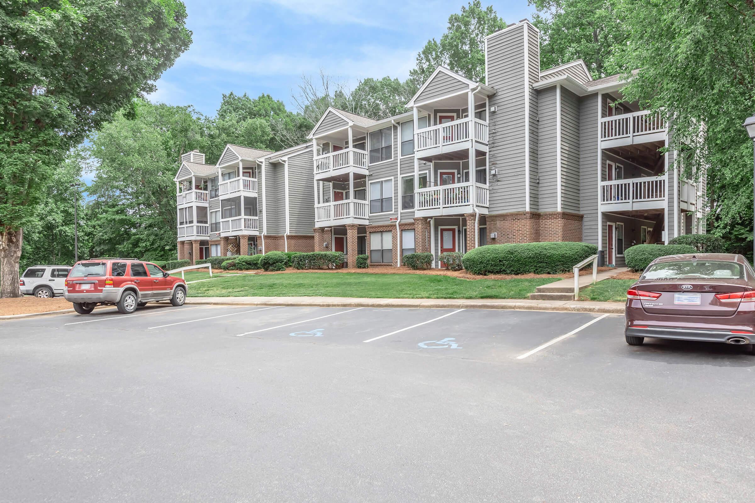 Exterior view of a multi-unit apartment building surrounded by greenery. The building features multiple balconies and a mix of brick and siding. A parking lot in front includes two parked cars, with visible designated accessible parking spaces.