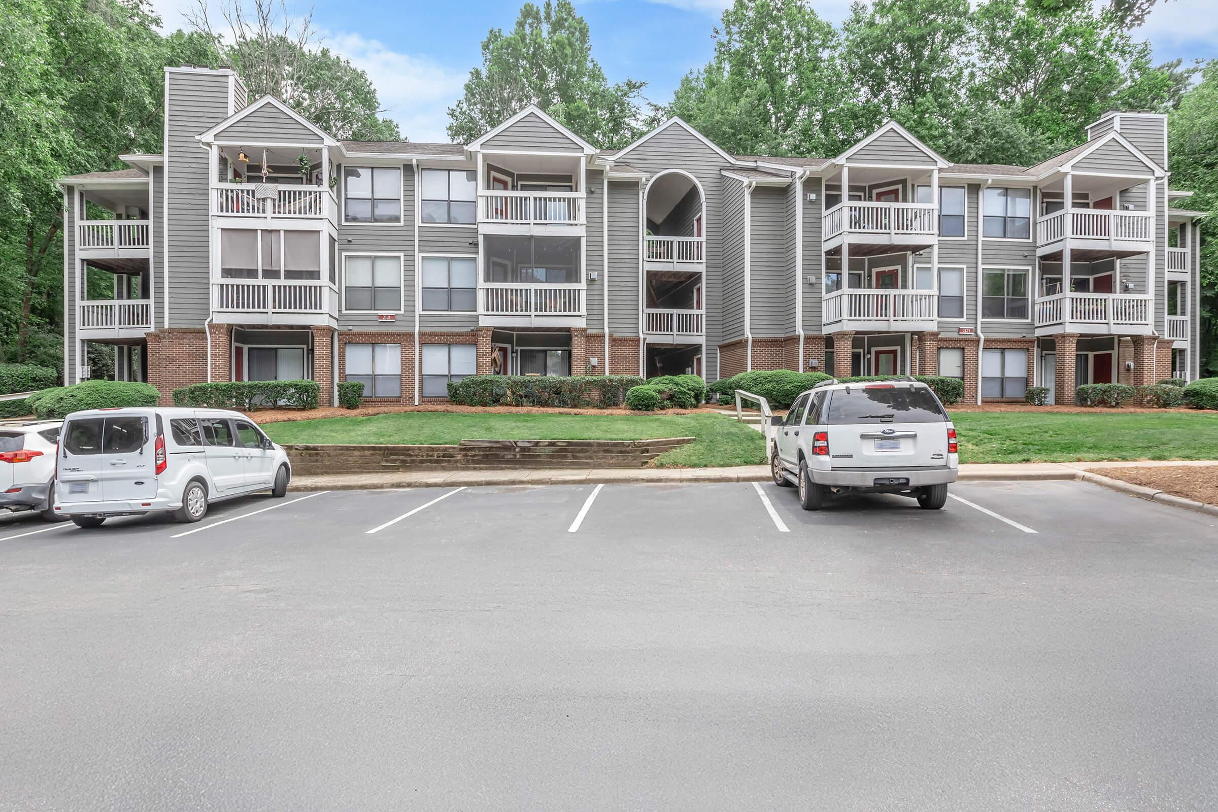 A multi-story apartment building surrounded by greenery, with balconies on each unit. Several parked vehicles are in the foreground, with a well-maintained lawn and trees in the background under a partly cloudy sky.