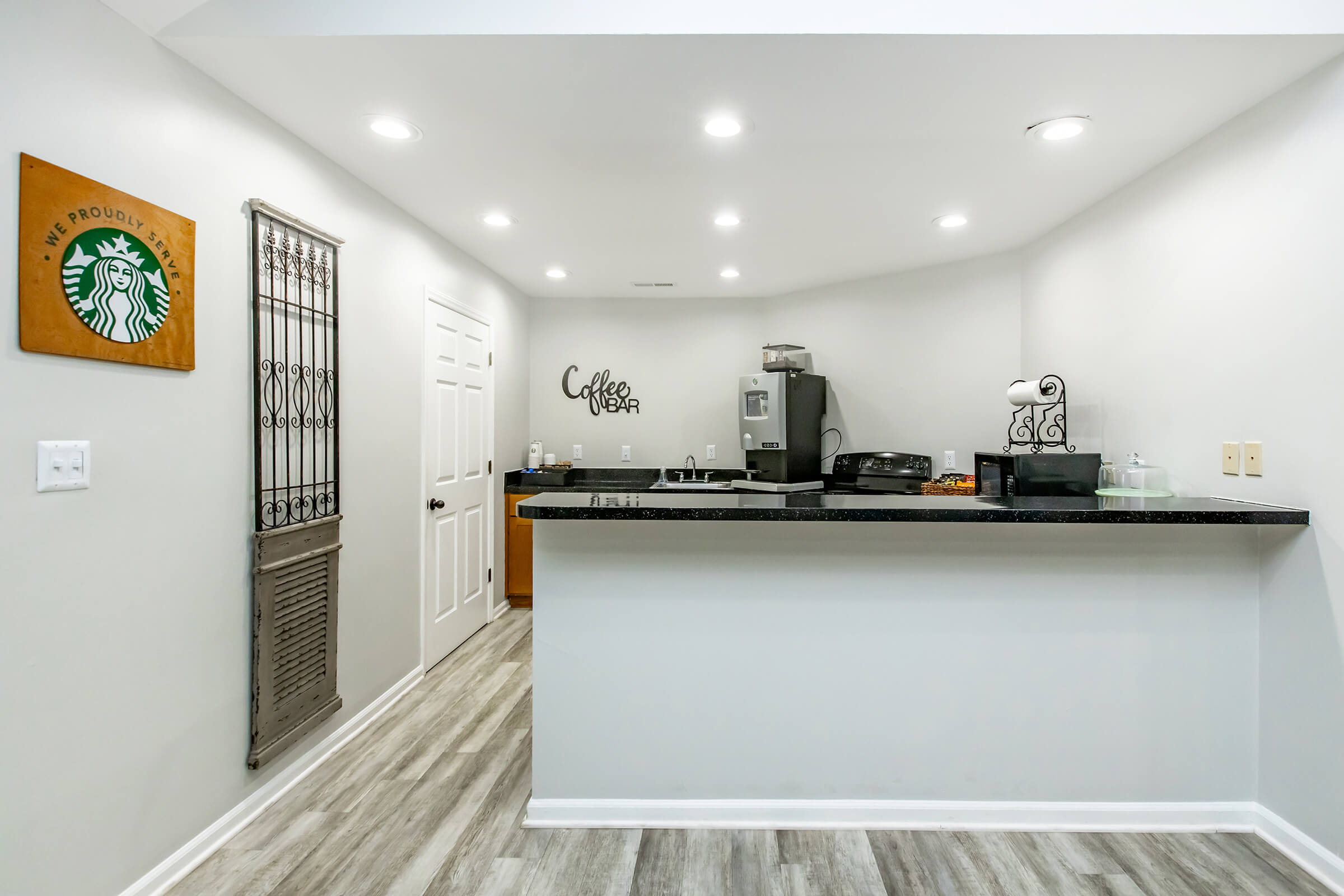 A modern kitchen space featuring a black counter, white walls, and wooden flooring. The kitchen includes a coffee bar sign, a stainless steel refrigerator, and a decorative Starbucks sign on the wall. There is also a metal decorative piece on the wall and various kitchen items on the counter.