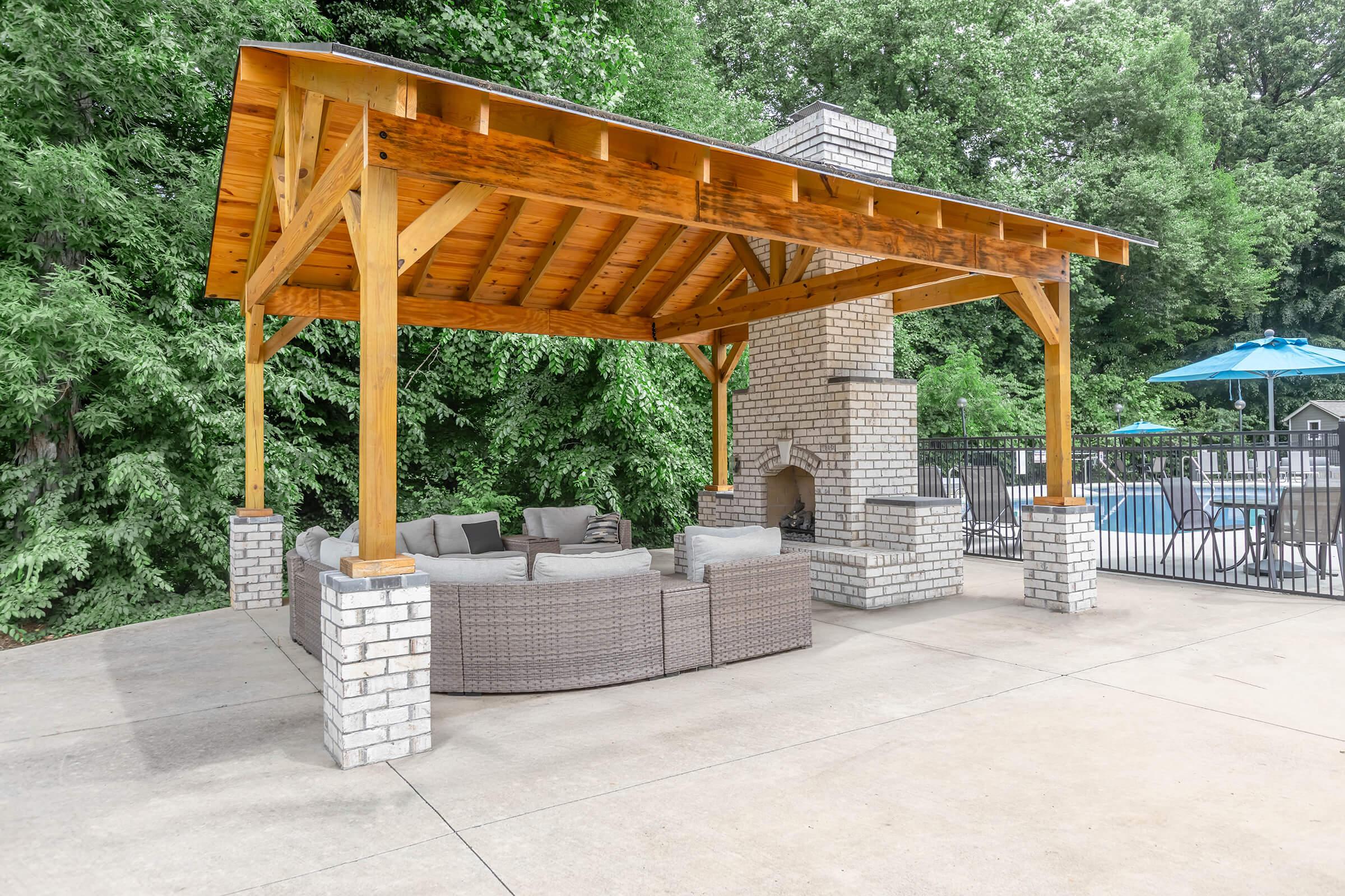 A wooden gazebo with a slanted roof and stone fireplace is set in a landscaped outdoor area. Inside the gazebo, there is a circular seating arrangement made of wicker, surrounded by greenery. In the background, a swimming pool is visible, along with an umbrella for shade.