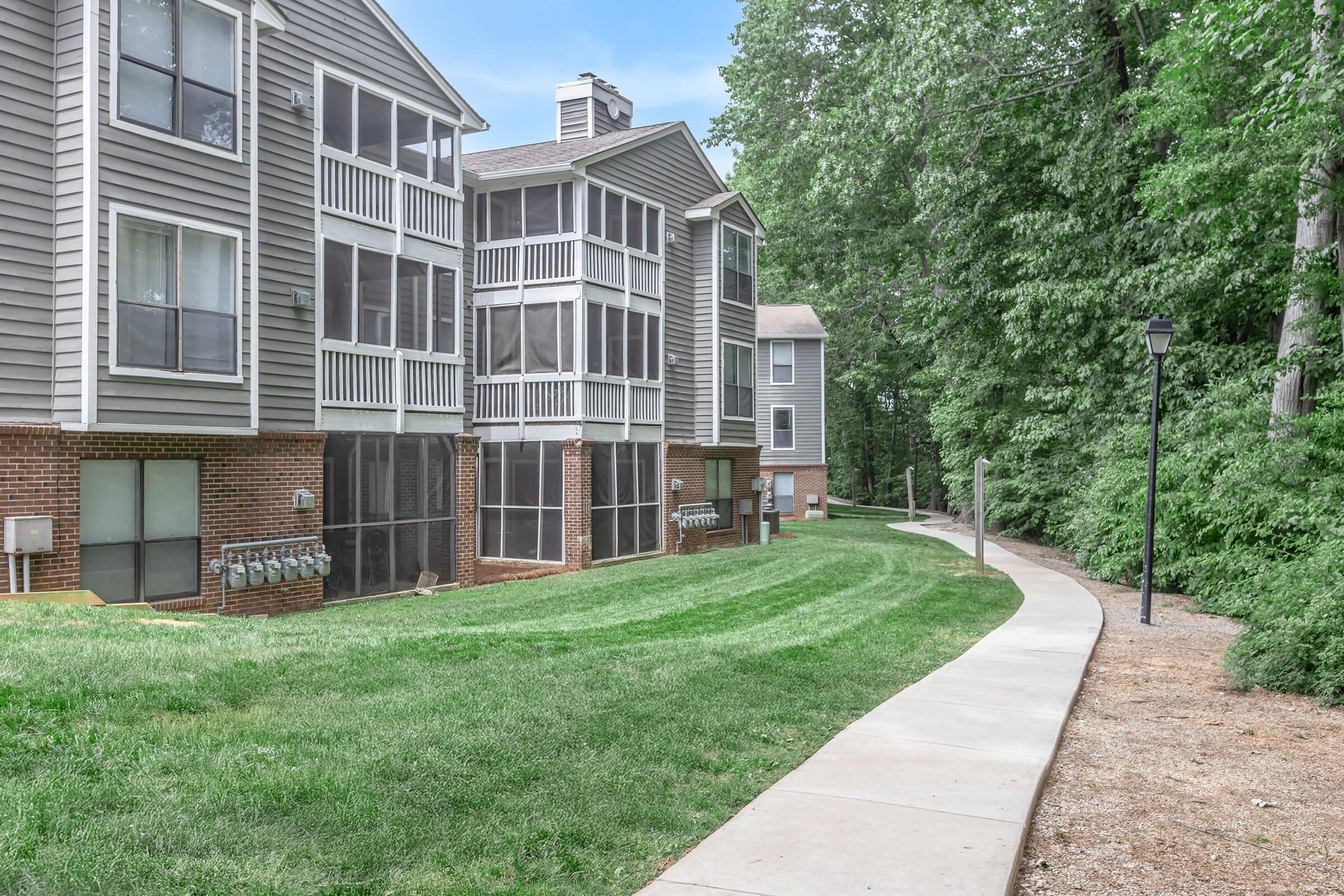 View of a landscaped pathway alongside two multi-story apartment buildings with screened-in patios. Lush green grass and trees flank the walkway, providing a tranquil setting. Mailboxes are visible near the building's entrance, emphasizing a residential atmosphere.