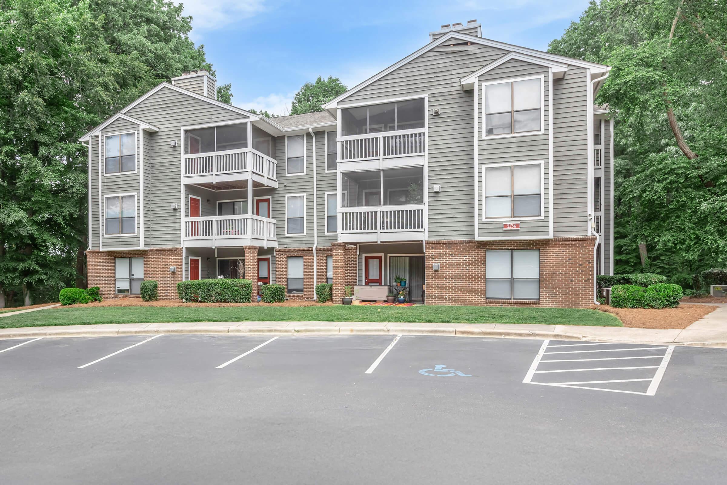 A multi-story apartment building with a gray exterior and white balconies. Green bushes line the front, and a parking lot is visible in the foreground, featuring designated spots, including one for handicapped parking. Trees provide a natural backdrop in the background.