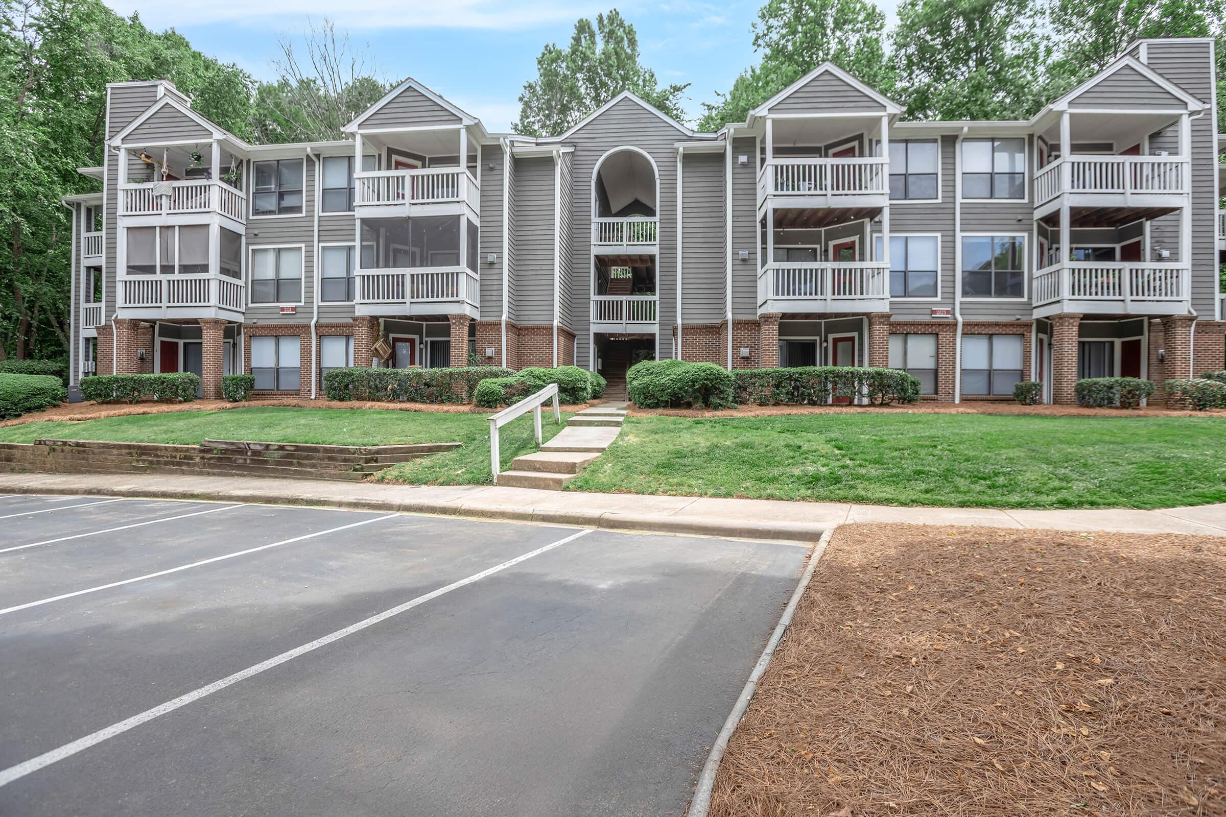 A multi-unit apartment building with three floors, featuring balconies and large windows. The building has a grey exterior with brick accents, surrounded by lush greenery and manicured lawn. In the foreground, a parking lot with marked spaces. Clear blue sky in the background.