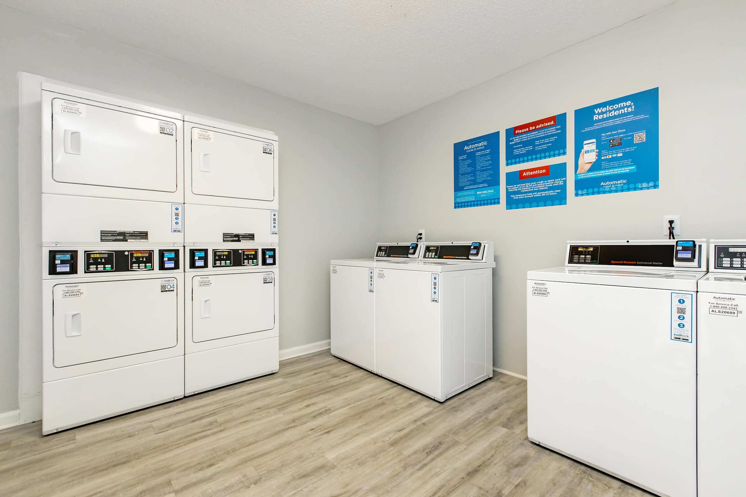 A clean laundry room featuring multiple washing machines and dryers. The machines are white and modern, placed against a light gray wall. Informational posters hang on the wall, providing instructions and guidelines for using the facilities. The flooring is light-colored, enhancing the bright and tidy atmosphere.